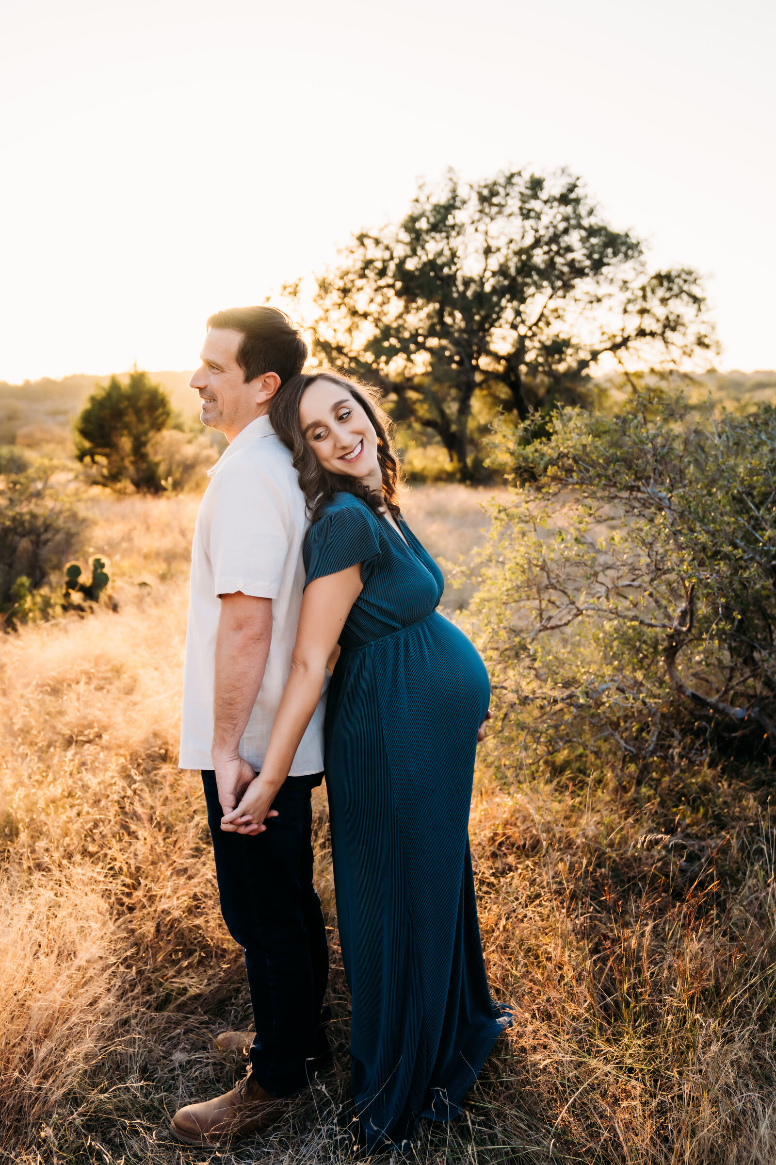 Maternity Photographer, husband and wife stand back-to-back and hold hands, mom is expecting, they are in a dry field