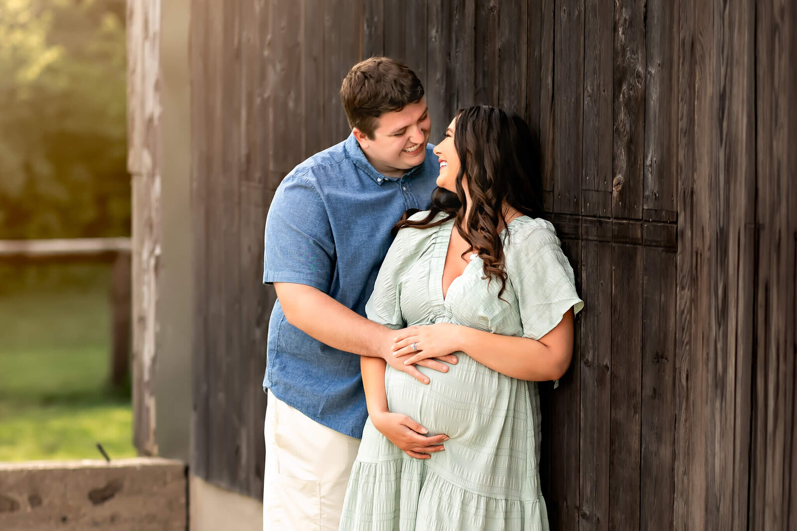 A smiling couple stands close together by a wooden wall. The woman, in a light green dress, cradles her pregnant belly as the man embraces her from behind. They gaze at each other lovingly, capturing a tender moment of pregnancy photography.