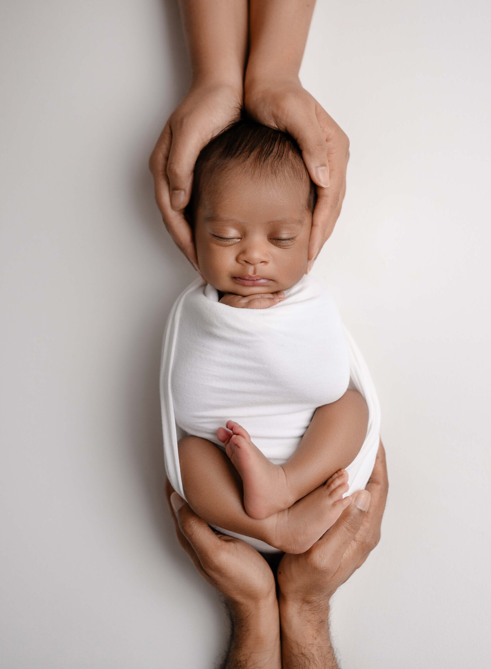 Newborn baby in white swaddled with Mom cradling his head and Dad cradling his bottom. Newborn Photography session photographed at Jenni Nikki's photo studio in Marietta, GA.