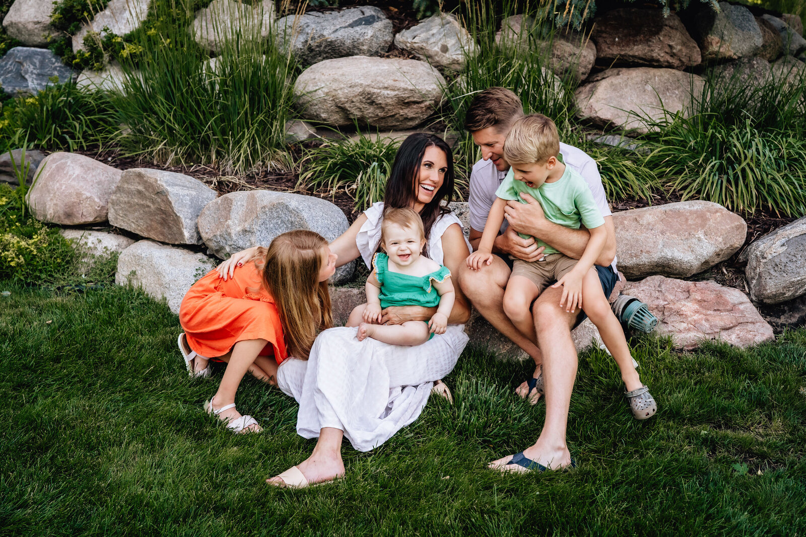A family of five sits on a grassy lawn in front of a rock wall, with the parents smiling and engaging with their three young children.