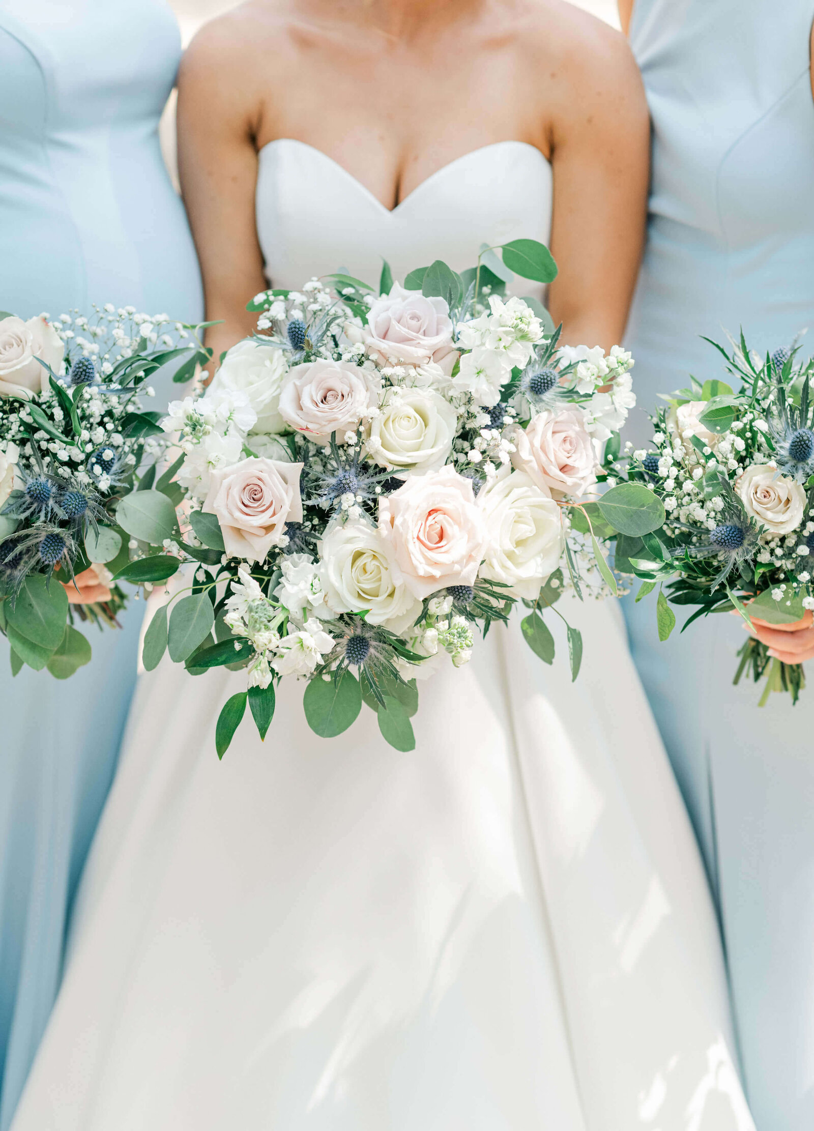 Bride holding wedding bouquet featuring white and pastel colored roses with eucalyptus against brides white dress and bridesmaids powder blue dress
