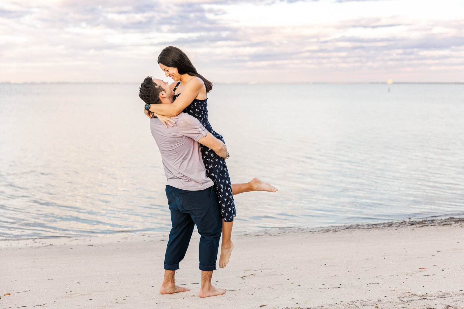 A husband lifts his wife in the air as they stand on a beach with the ocean in background