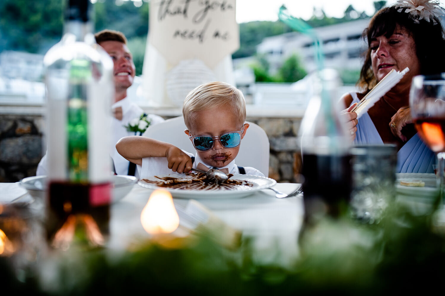 Flower boy in sunglasses eating cake with choclate all over his mouth during the wedding speeches