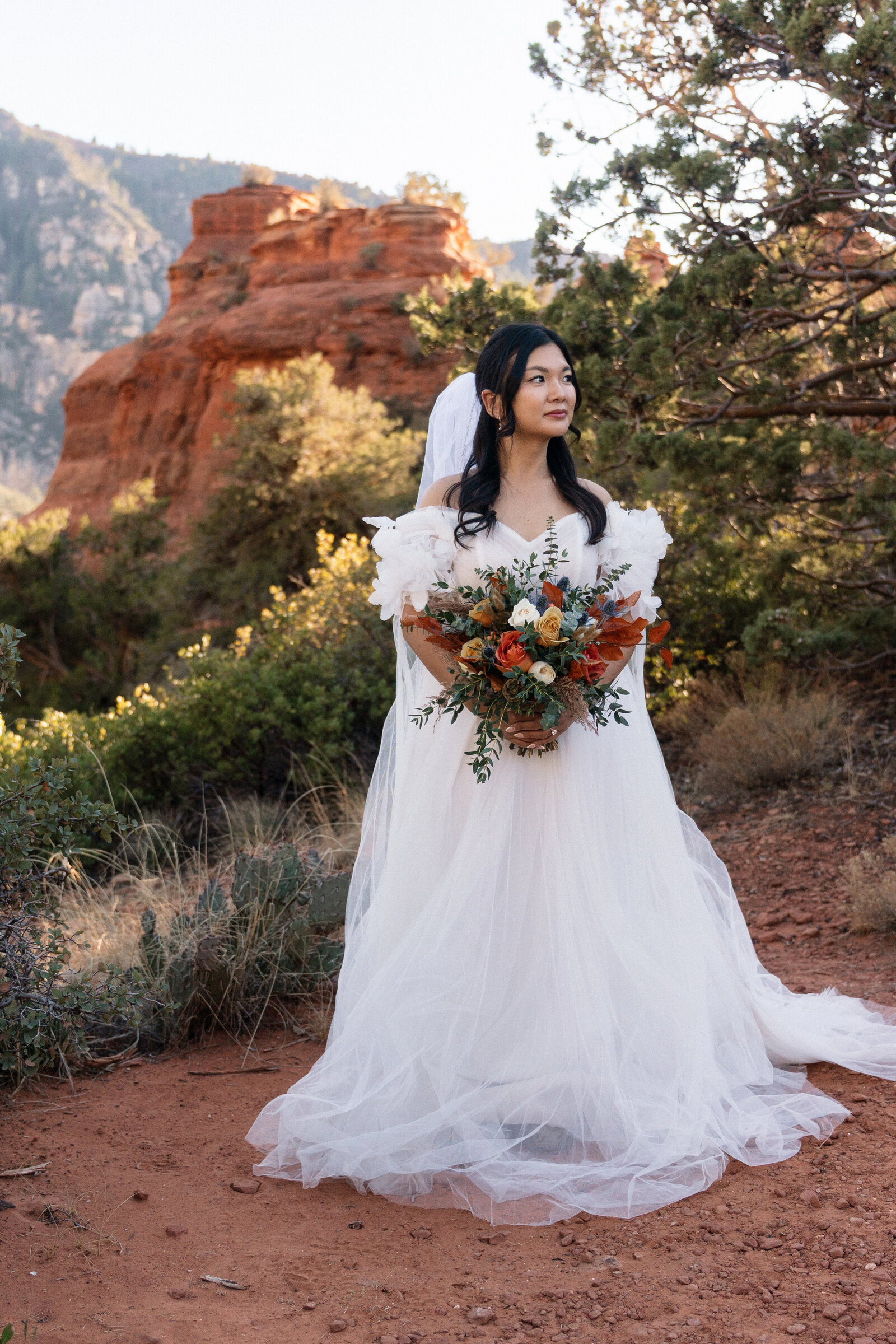 Bride in a flowing off-the-shoulder wedding dress holding a vibrant bouquet during her elopement at Merry Go Round Rock in Sedona, Arizona, with red rock formations and desert foliage in the background.