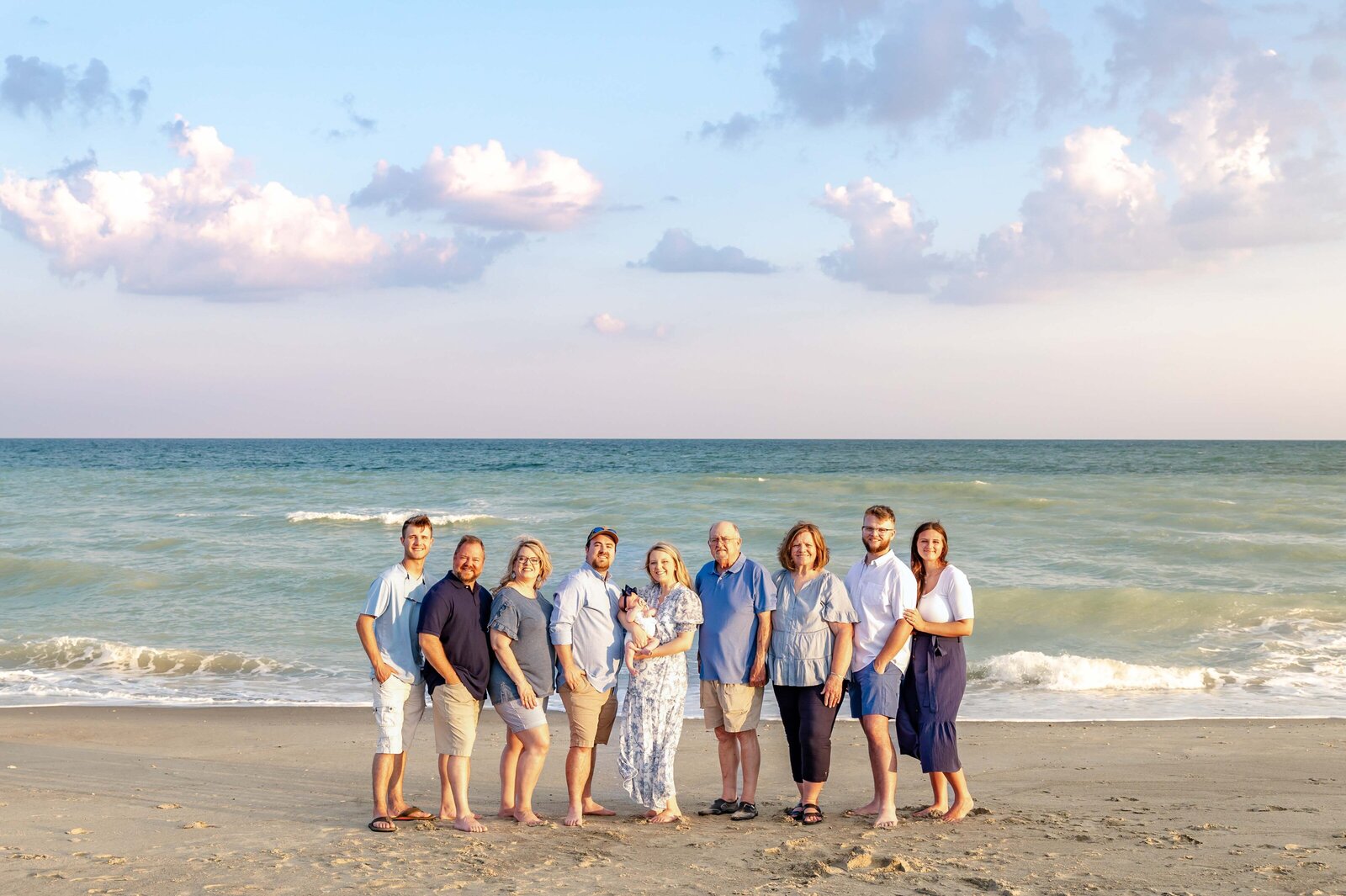 A family poses on the beach at sunset, with soft clouds and the ocean creating a serene backdrop. Dressed in blue and white, they capture a relaxed coastal moment, perfect for North Carolina family photography, highlighting beachside elegance.