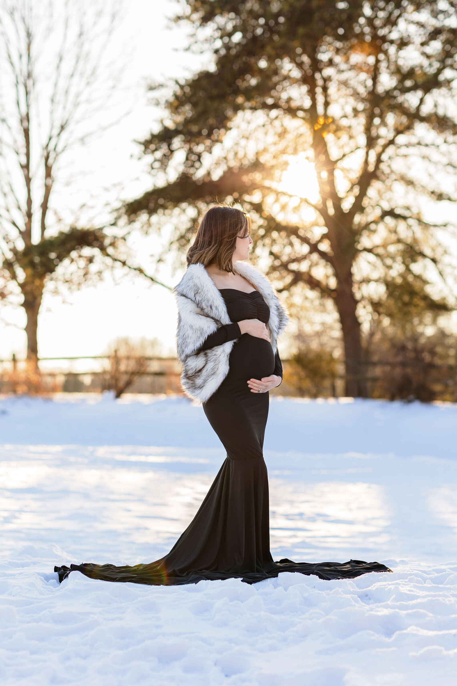 A mother-to-be standing in the snow in a black dress and fur shrug at sunset.