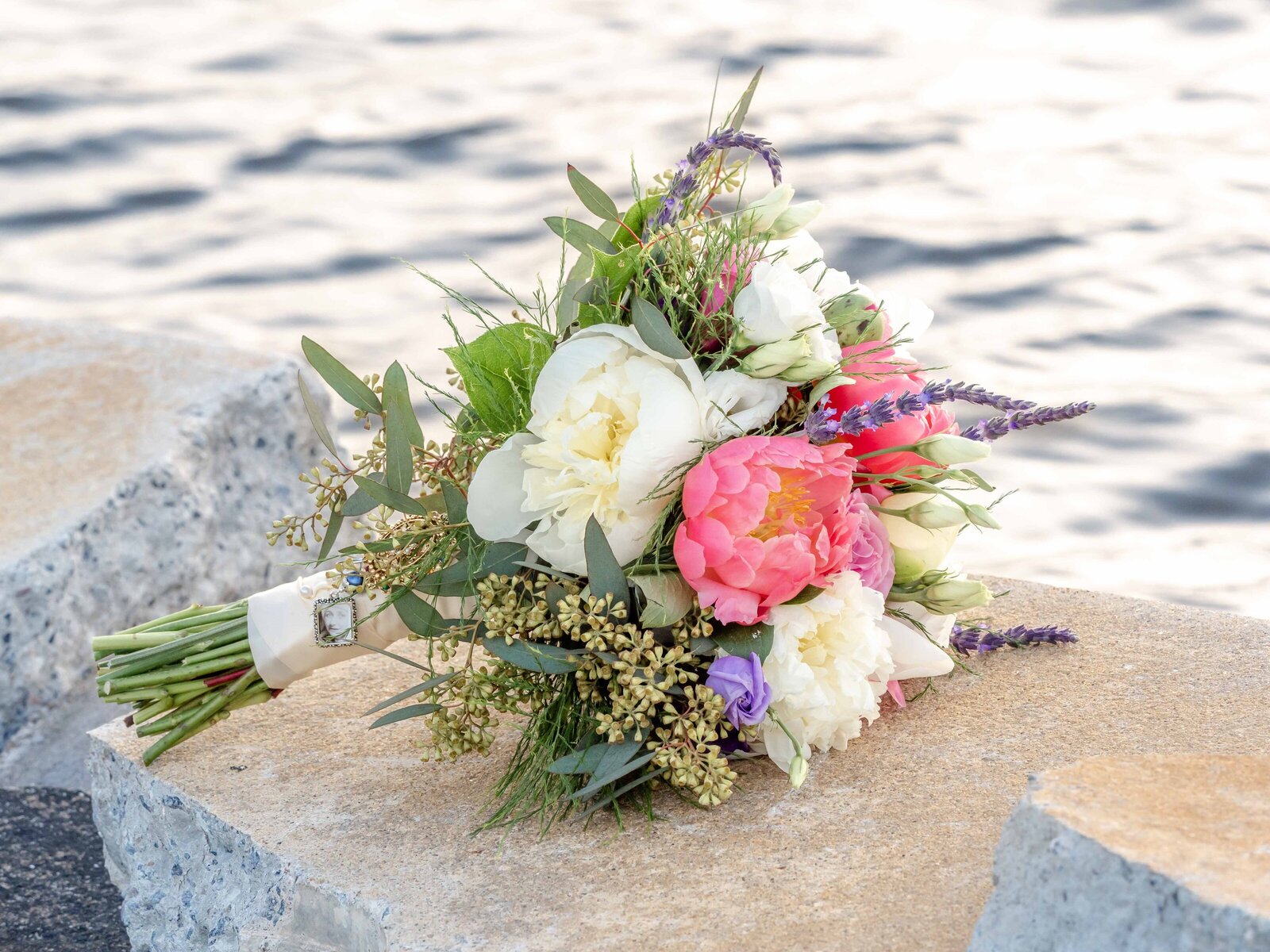A vibrant wedding bouquet with pink peonies, white roses, lavender, and greenery rests on a stone ledge by the water, captured during a Wrightsville Beach, NC wedding, blending natural beauty and colorful elegance.