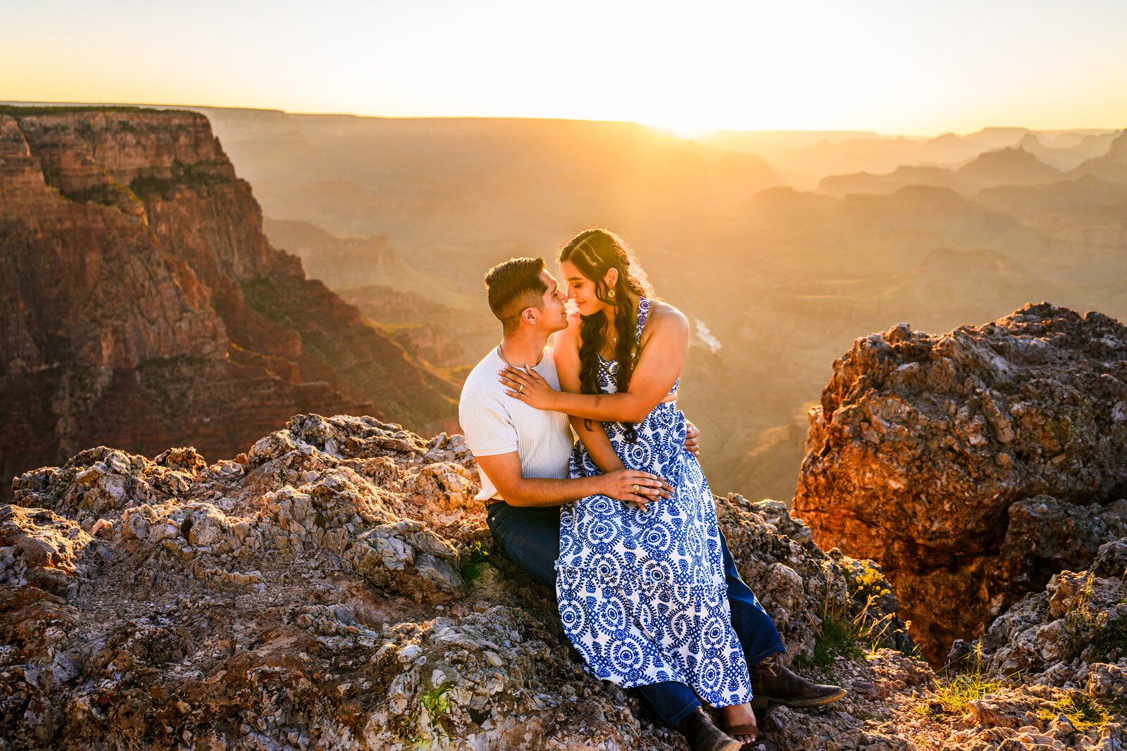 Engaged couple at Grand Canyon National Park holding each other she is sitting on his lap with golden hour sunset behind them fading into canyon