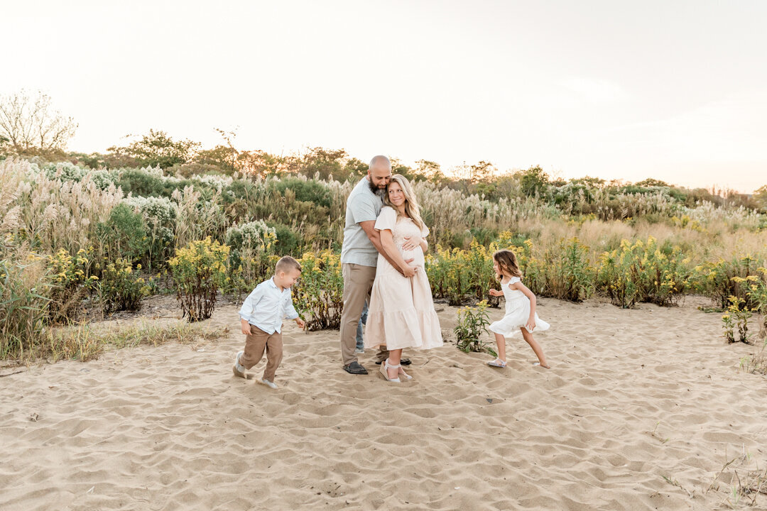 Young kids running around parents as they embrace duringFamily photo session on beach in NJ