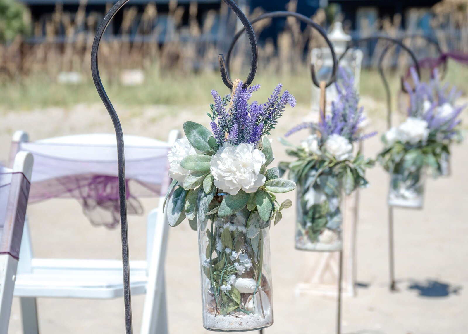 Elegant beach wedding aisle decor featuring lavender, white flowers, and greenery in hanging glass jars, captured at a Topsail Beach, NC wedding, blending rustic charm with coastal beauty.