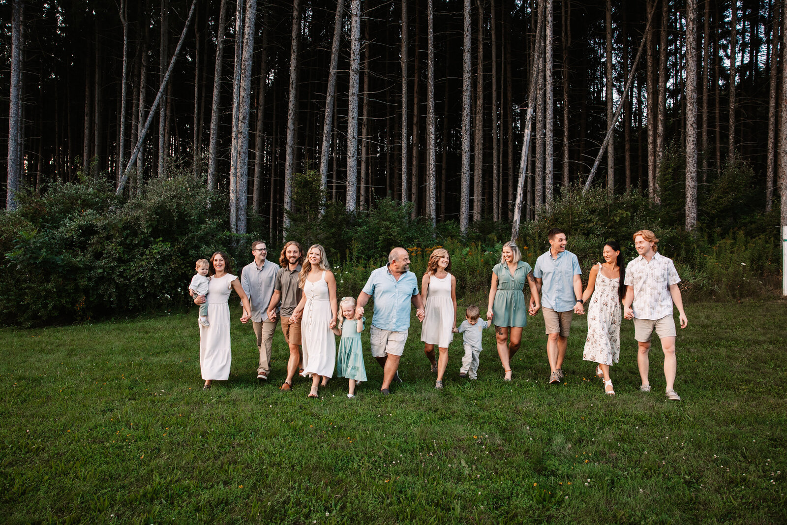 A large extended family walks in a field holding hands and smiling at each other as guided by a Milwaukee Family Photographer