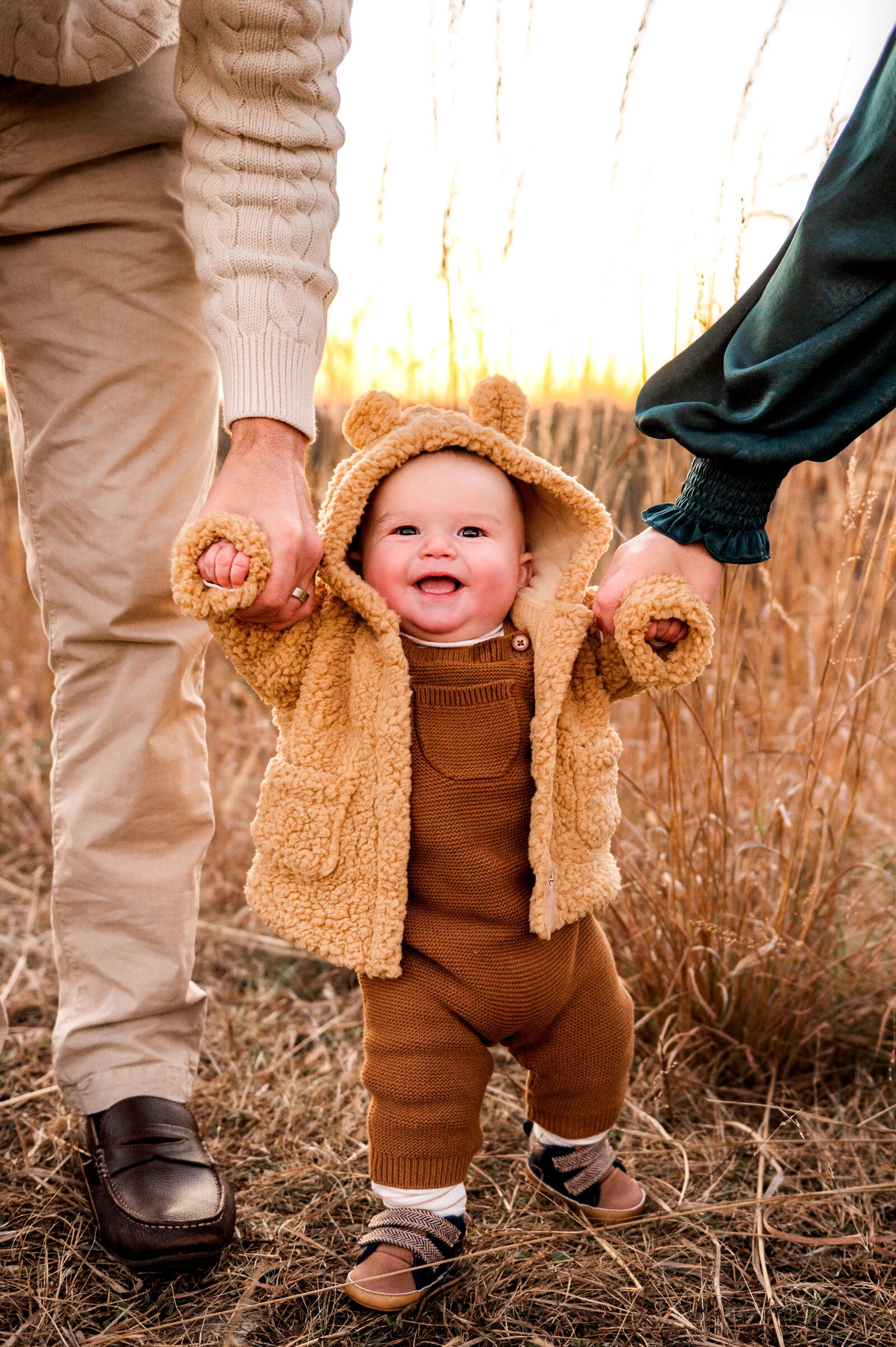 baby in a field at soldiers delight holding mom and dads hands as he smiles