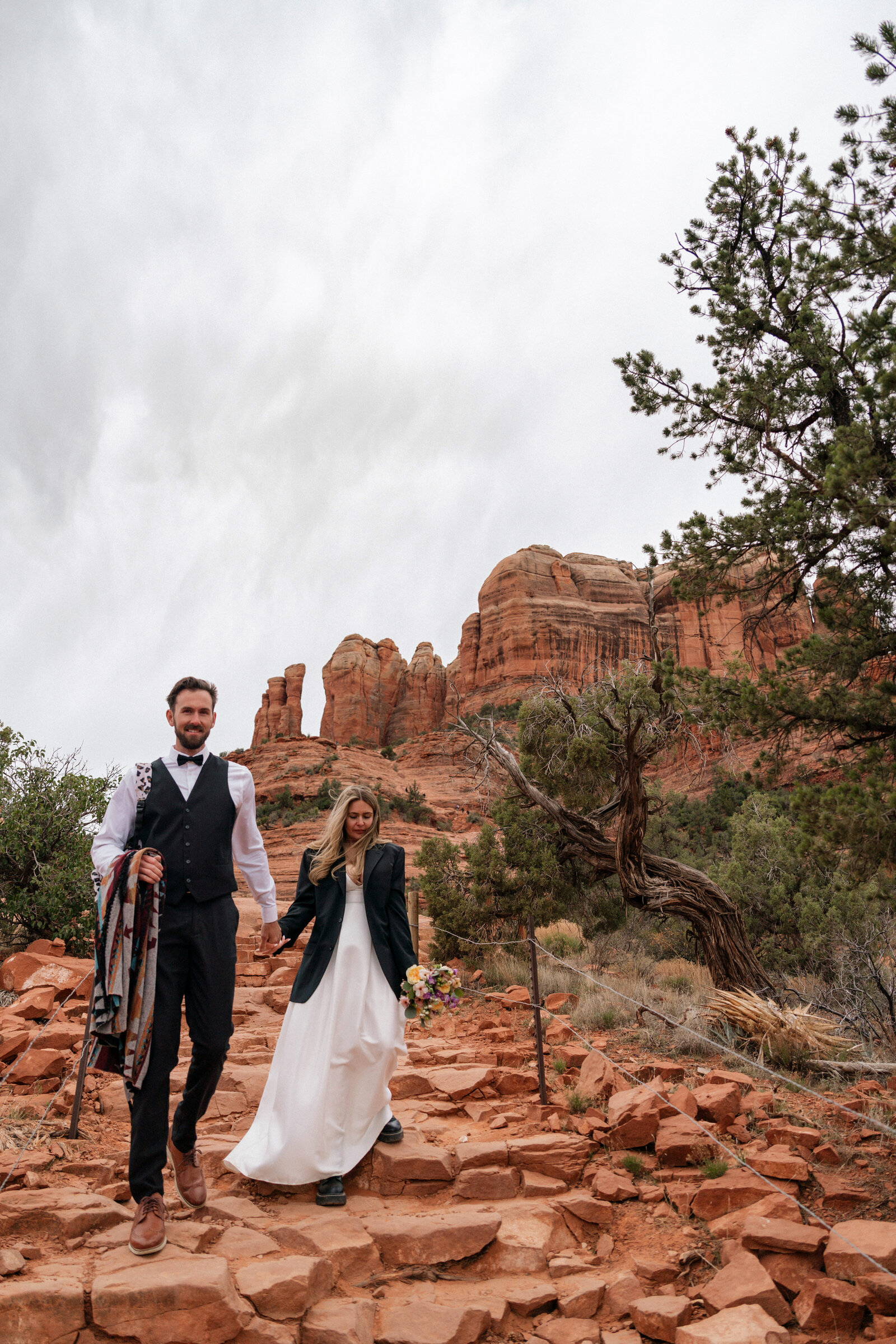 Bride and groom hiking down a rocky trail at Cathedral Rock in Sedona, Arizona, wearing wedding attire with jackets and boots, surrounded by red rock formations and desert trees on a cloudy day.