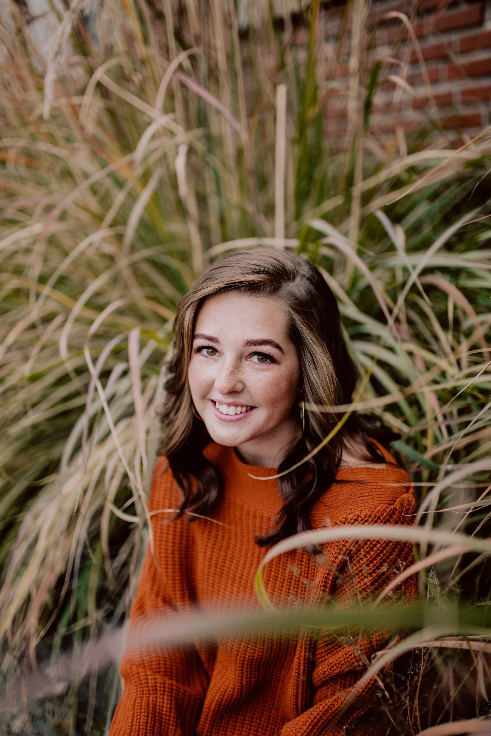 Girl taking senior photo, sitting next to tall green grass. She's smiling and looking at the camera.