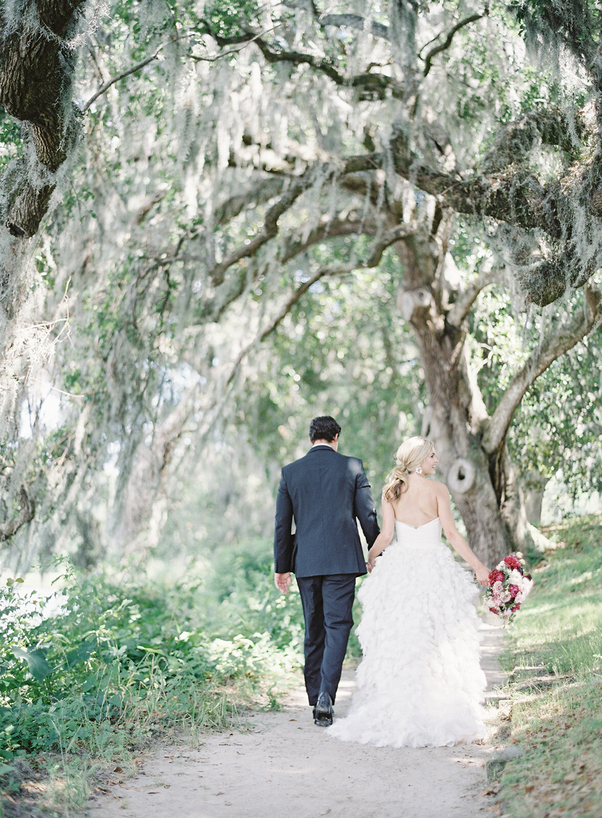 Couple under oak trees walking down path. Groom is in a black tux and bride is in a strapless gown with feathers as a skirt designed by Anne Barge. Photographed by wedding photographers in Charleston Amy Mulder Photography