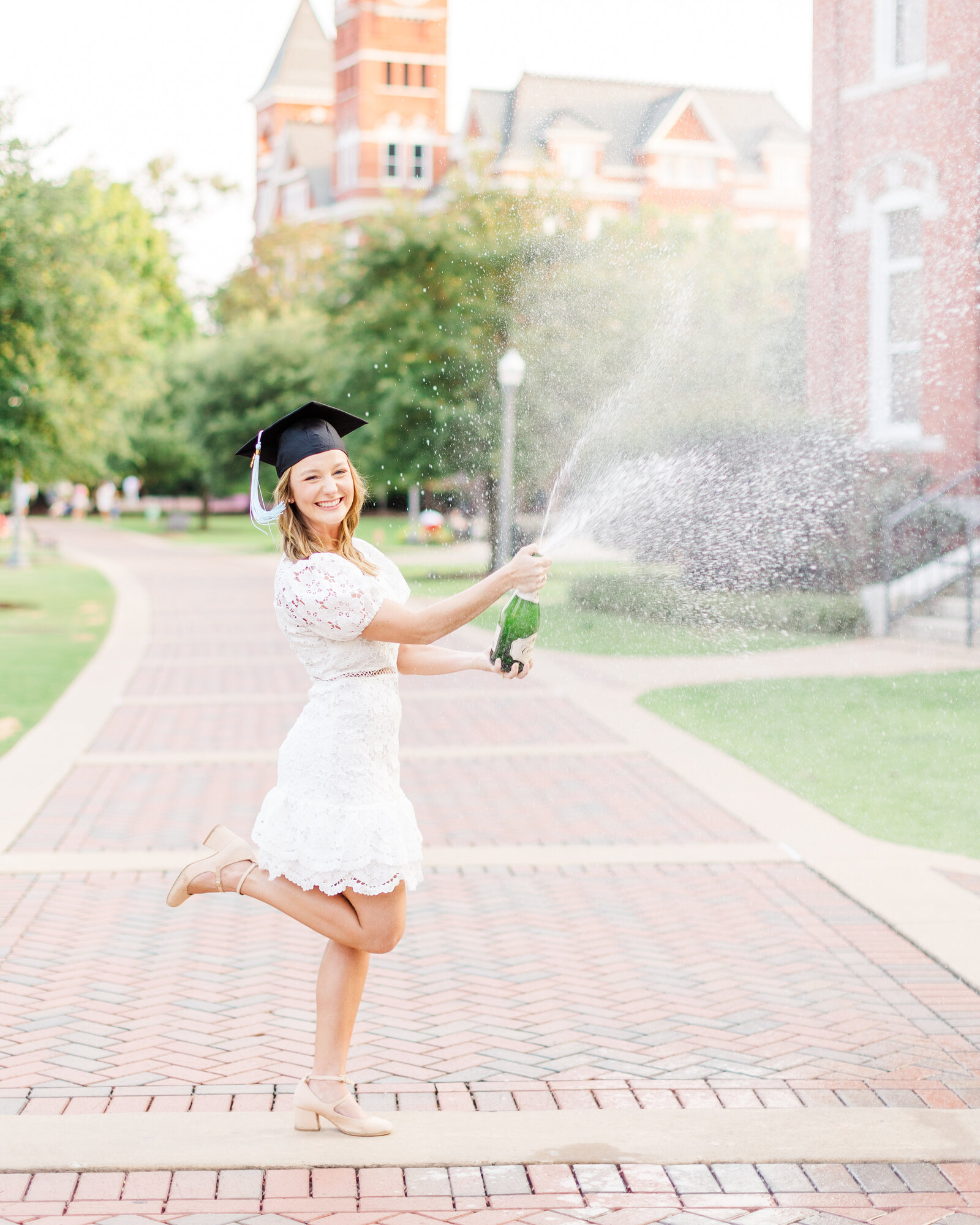 Shelby celebrating her graduation with a bottle of champagne, wearing a white lace dress and cap, captured during her senior photoshoot.