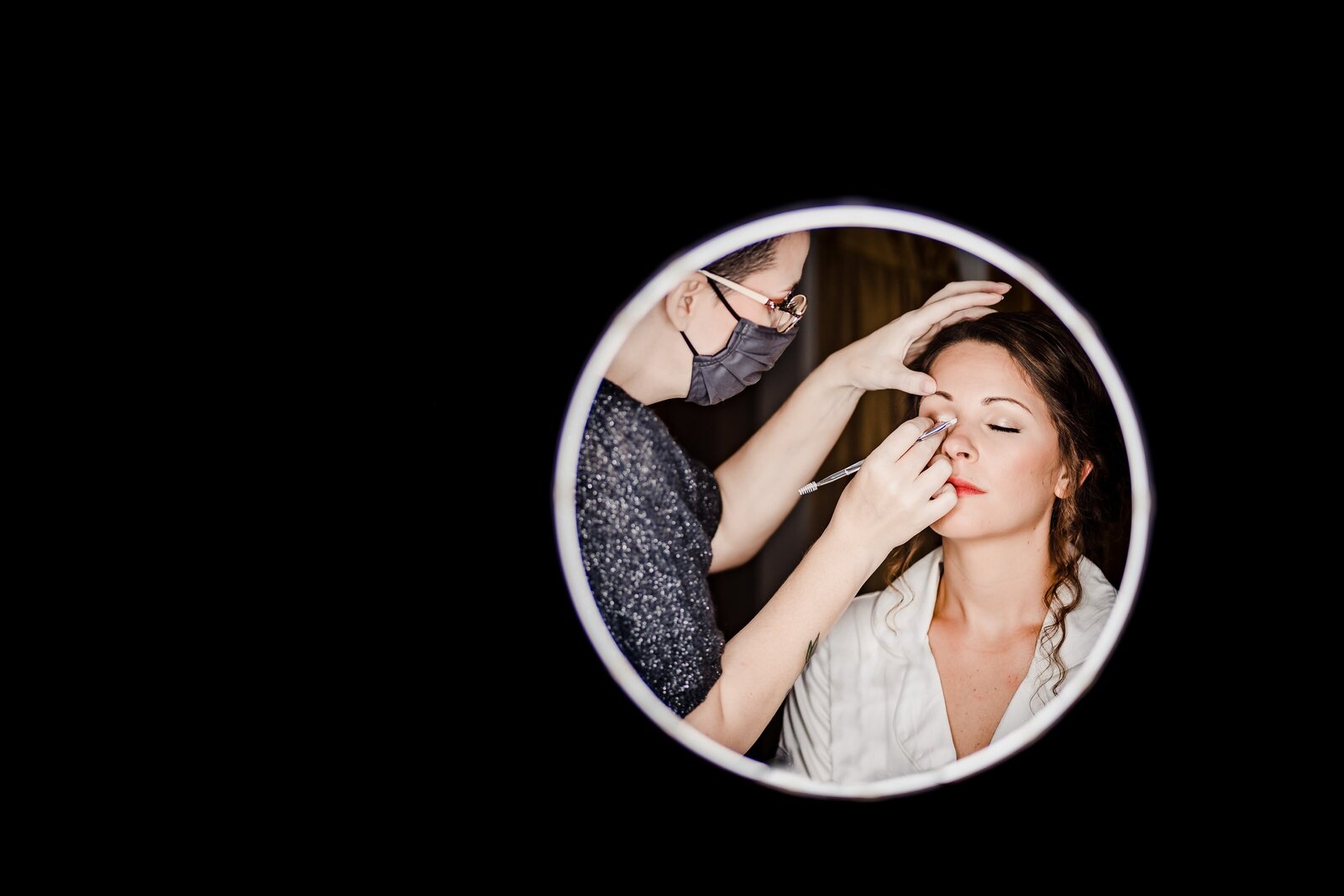 Creative wedding photography of a bride having her makeup done. She is seen through a ring light, while the rest of the frame is pitch black