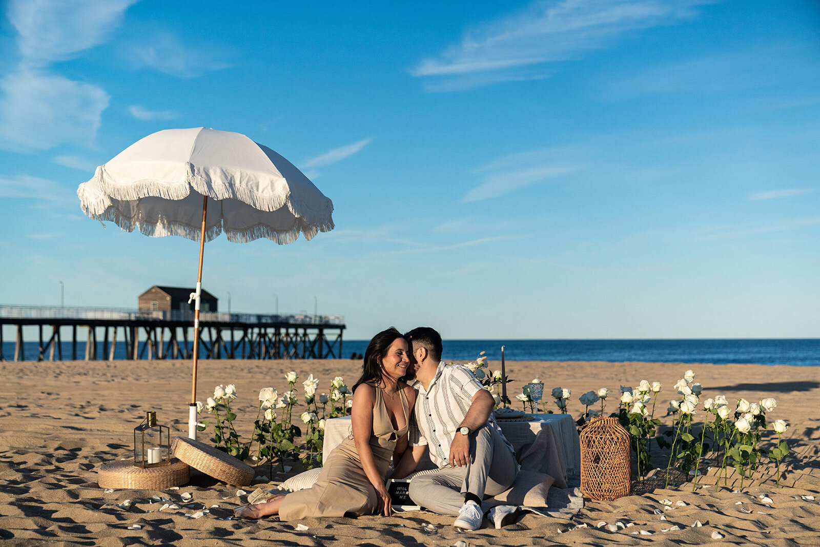 Couple sharing a kiss during a romantic beach picnic with white roses, a fringe umbrella, and ocean views, styled by Yours Truly, a wedding planner and designer in New Jersey