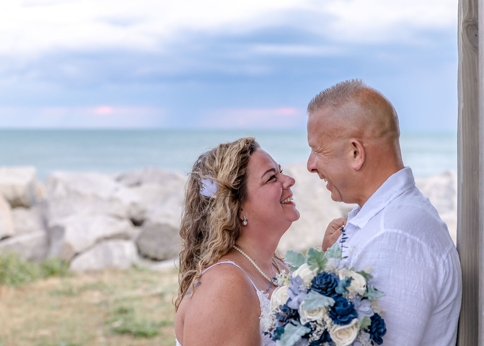 The bride and groom share a joyful moment under a beachside pavilion with the ocean and rocky shoreline in the background, captured during an Fort Fisher, NC wedding, blending romance with coastal charm.