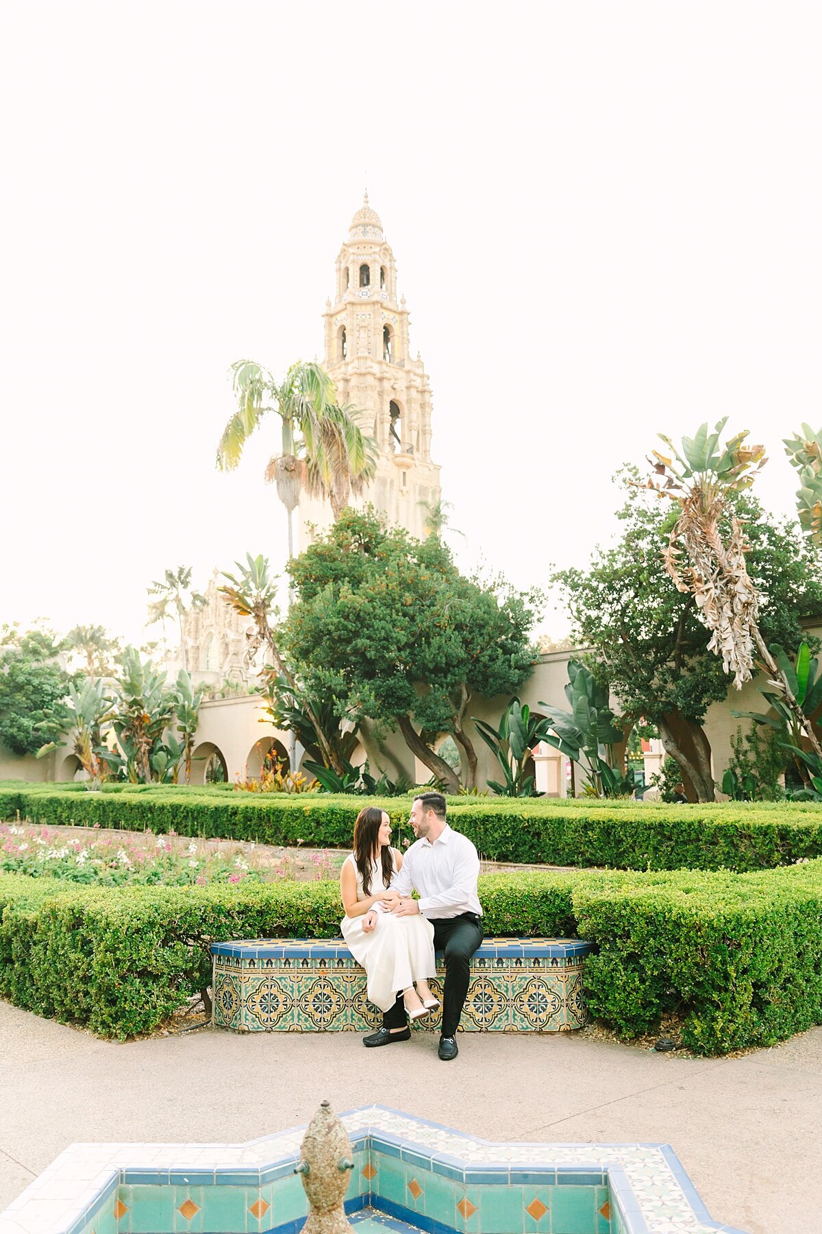Engaged couple sitting together in front of the tower at Balboa Park during sunset at their photography session.