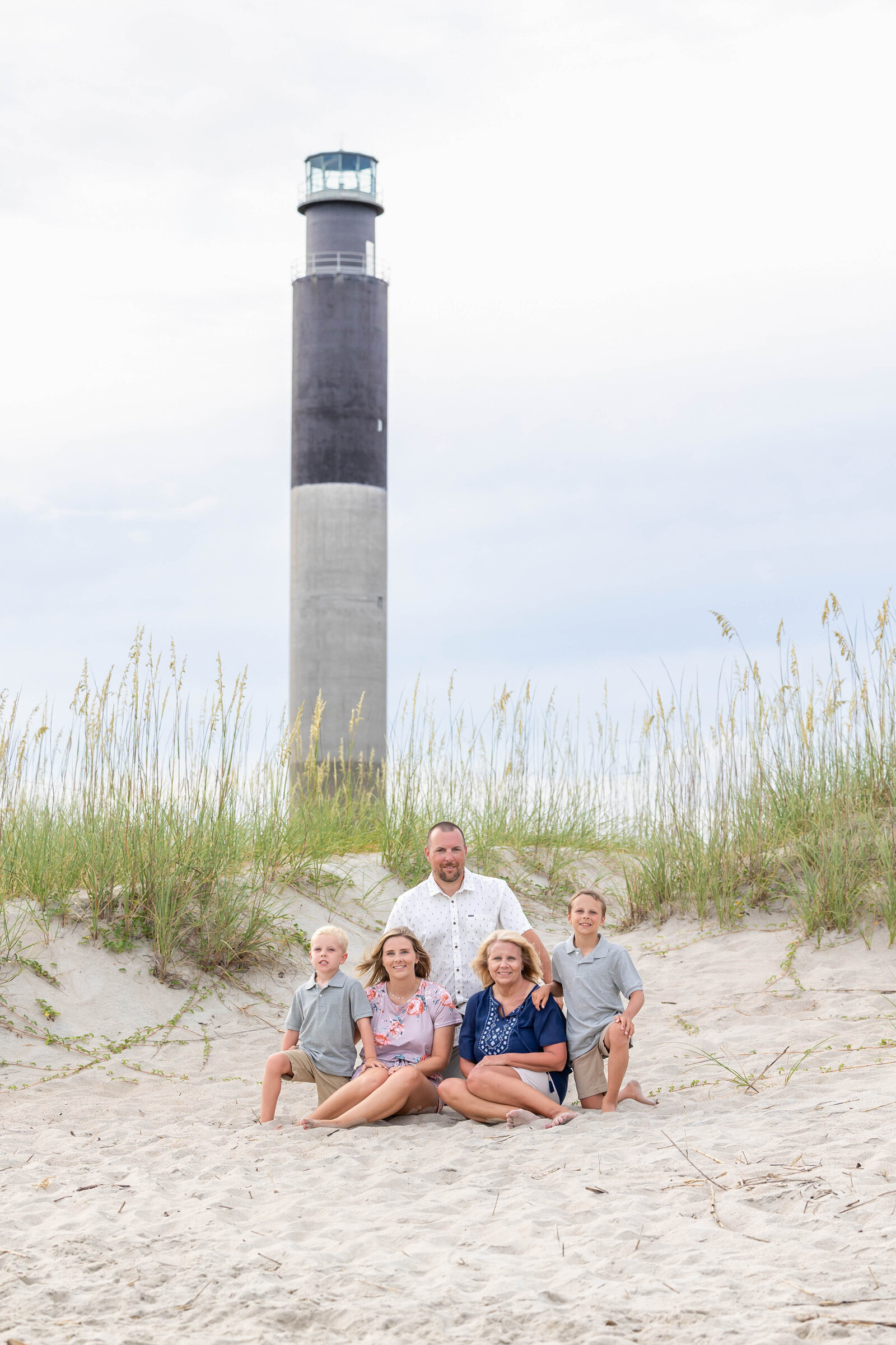 A family of five sits on the sandy dunes at Caswell Beach, with the iconic Oak Island Lighthouse towering in the background. The children and parents are dressed in casual beach attire, capturing a serene moment. Perfect for North Carolina family photography, highlighting scenic beach locations.