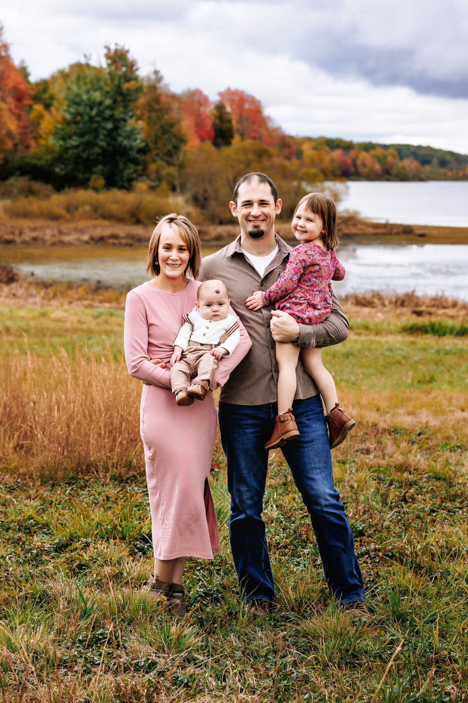 Family stands together for a portrait in front of  lake surrounded by beautiful fall foliage