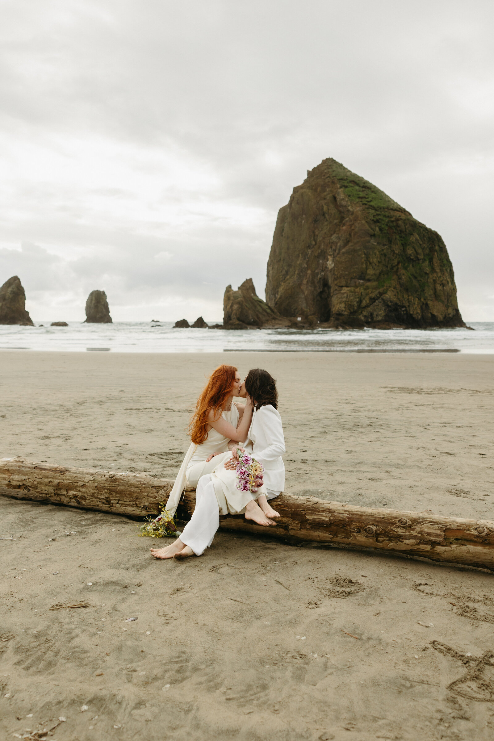 Two brides share a kiss on a log overlooking the Haystack rock at Cannon Beach.