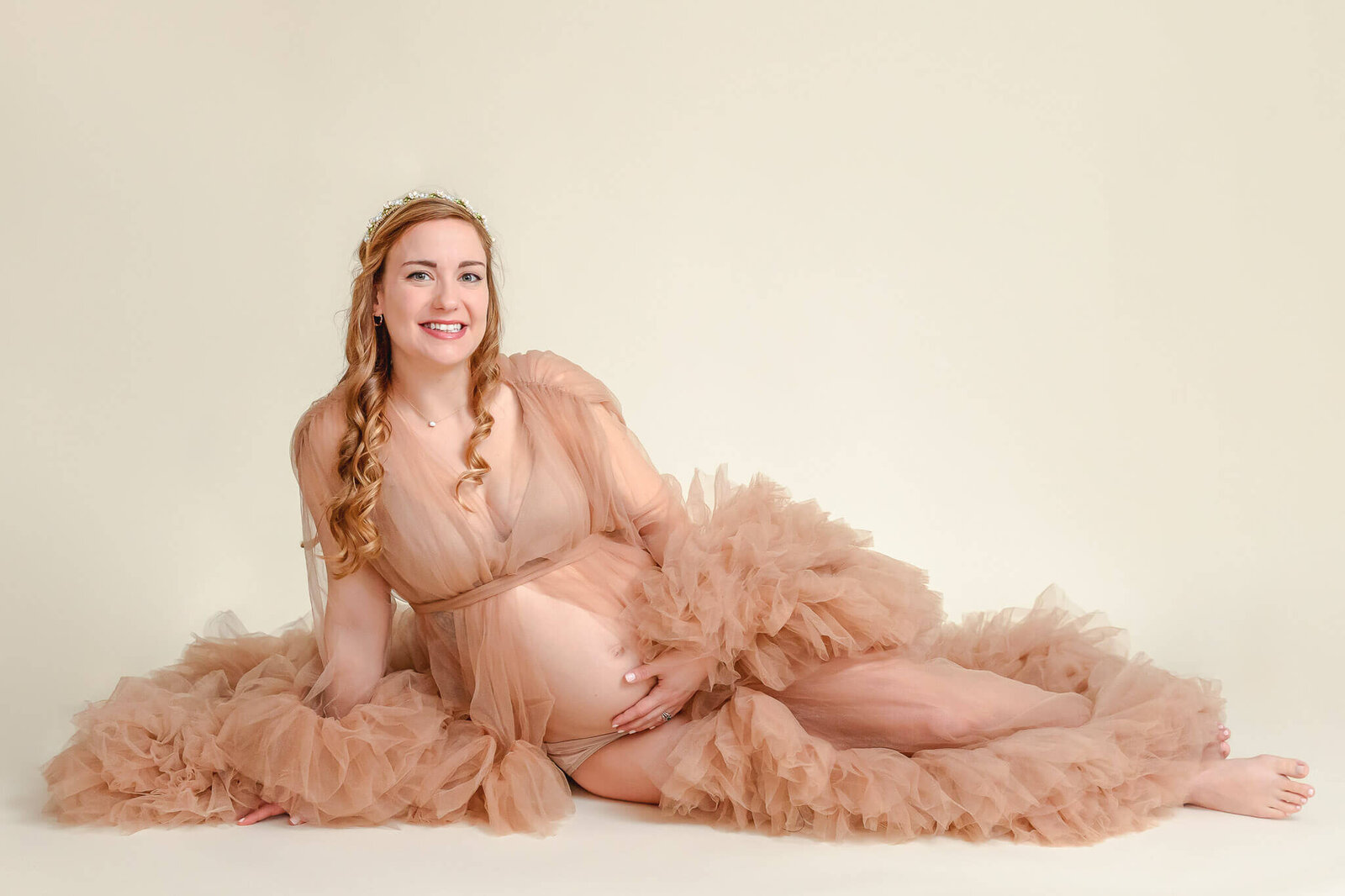 A mother-to-be in a tan robe sitting on the ground of a studio and holding her belly.