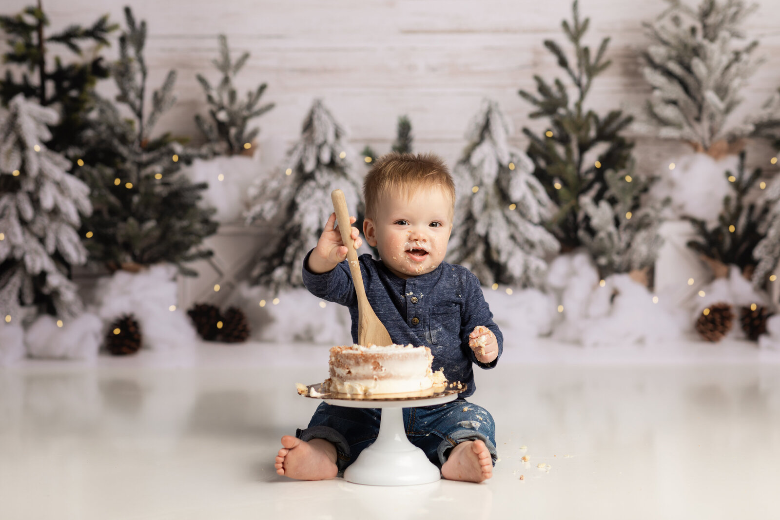 one year old boy in blue shirt holding spoon in cake for cake smash photoshoot