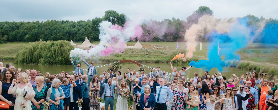 Large group of wedding guests smile at the camera for a group photo.  Colourful smoke flares are let off in the background showing plumes of orange, blue, pink and white smoke.  Hadsham Farm's lake and wedding tipi's can be seen in the background at this fun relaxed wedding