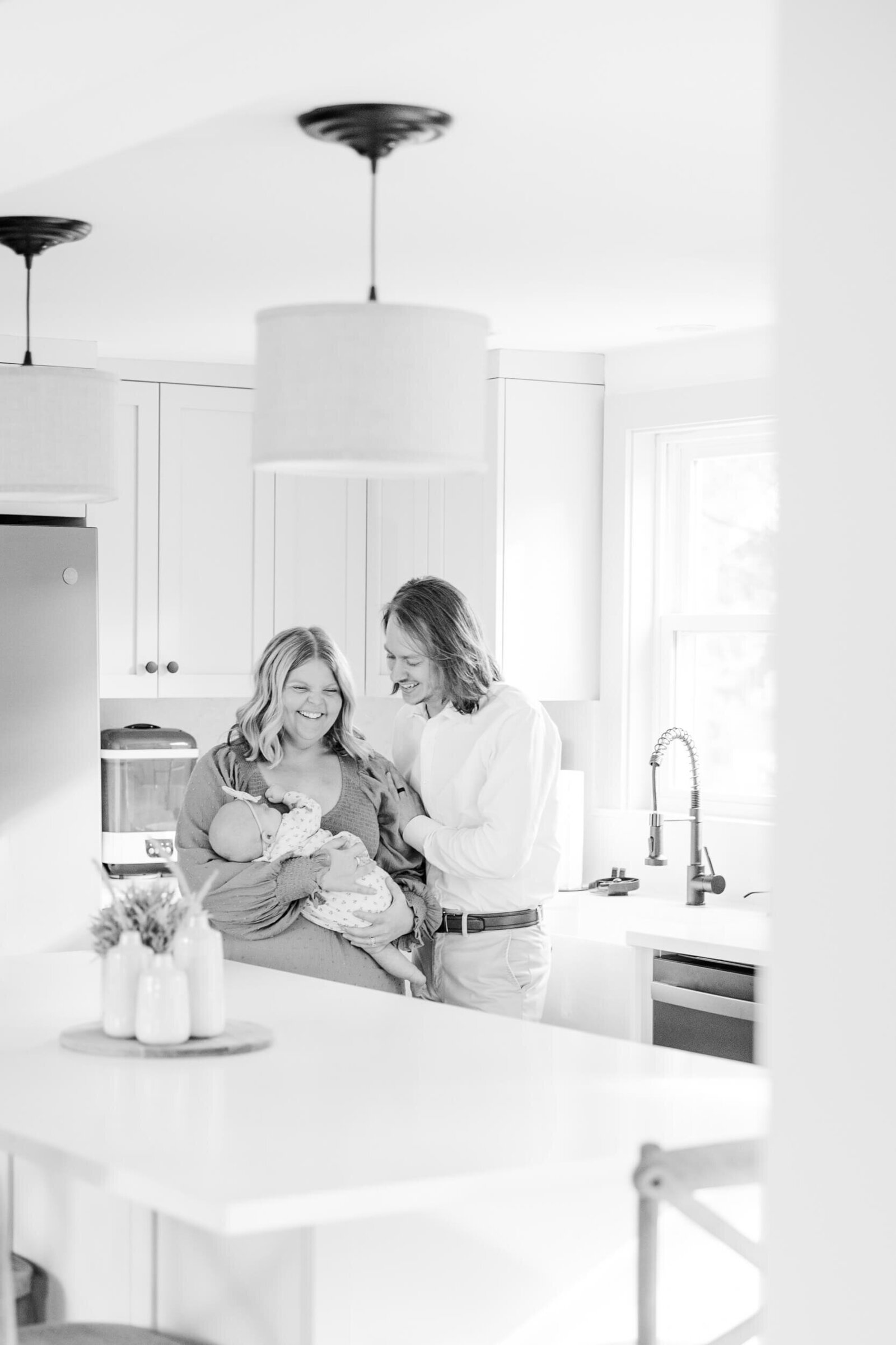 Black and white image of mom and dad talking in the kitchen while baby sleeps in mom's arms