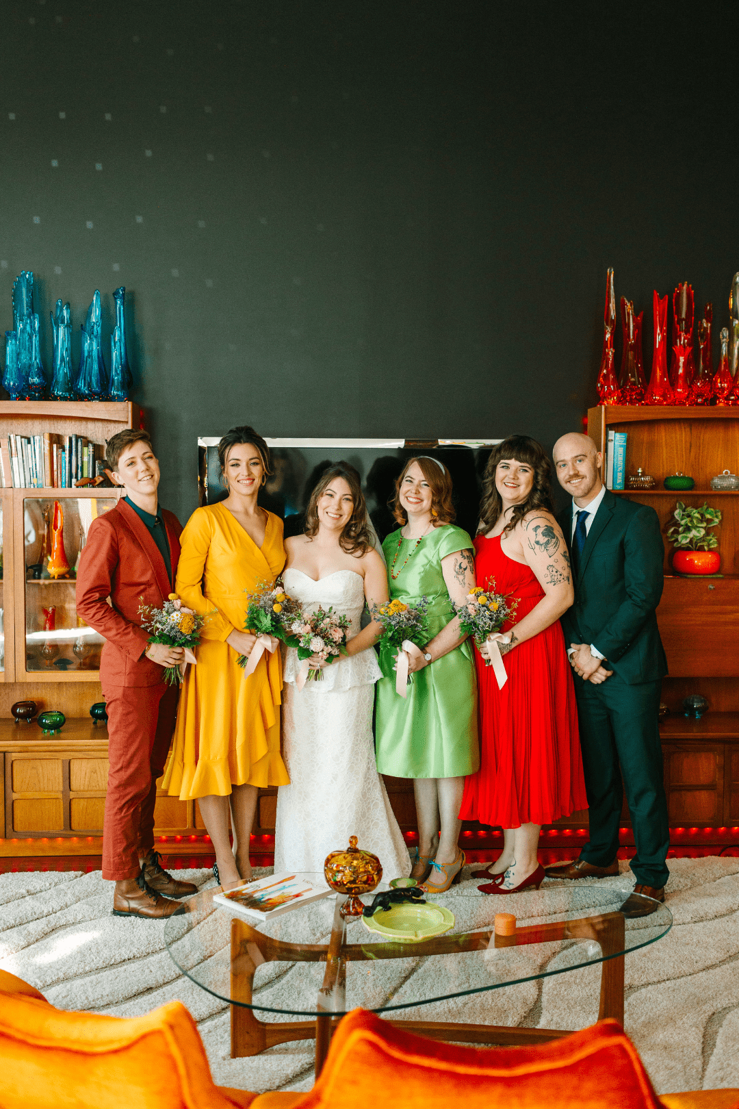 A bride and her 5 member wedding party  wearing colorful retro suits and dresses in front of a display of vintage glassware