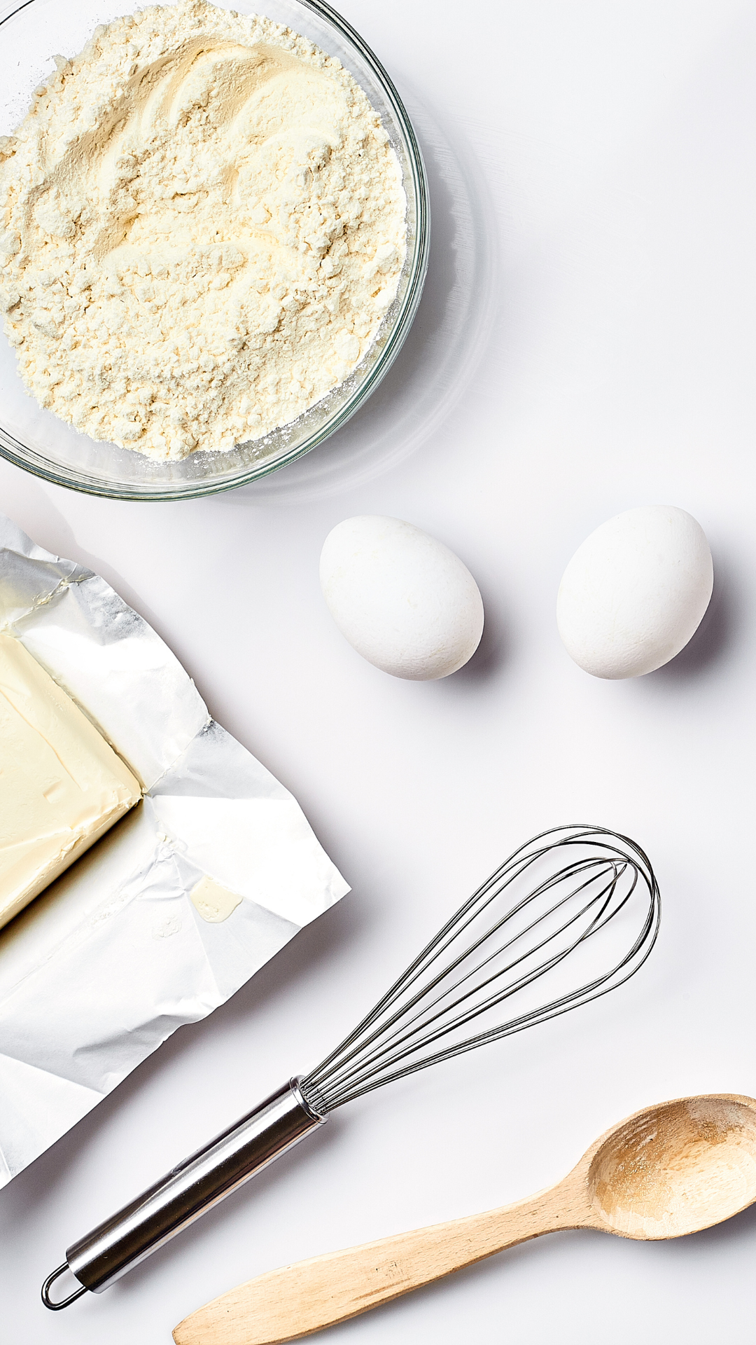 Basic baking ingredients laid out including flour, eggs, butter, a whisk, and a wooden spoon, ready for a healthy recipe preparation.