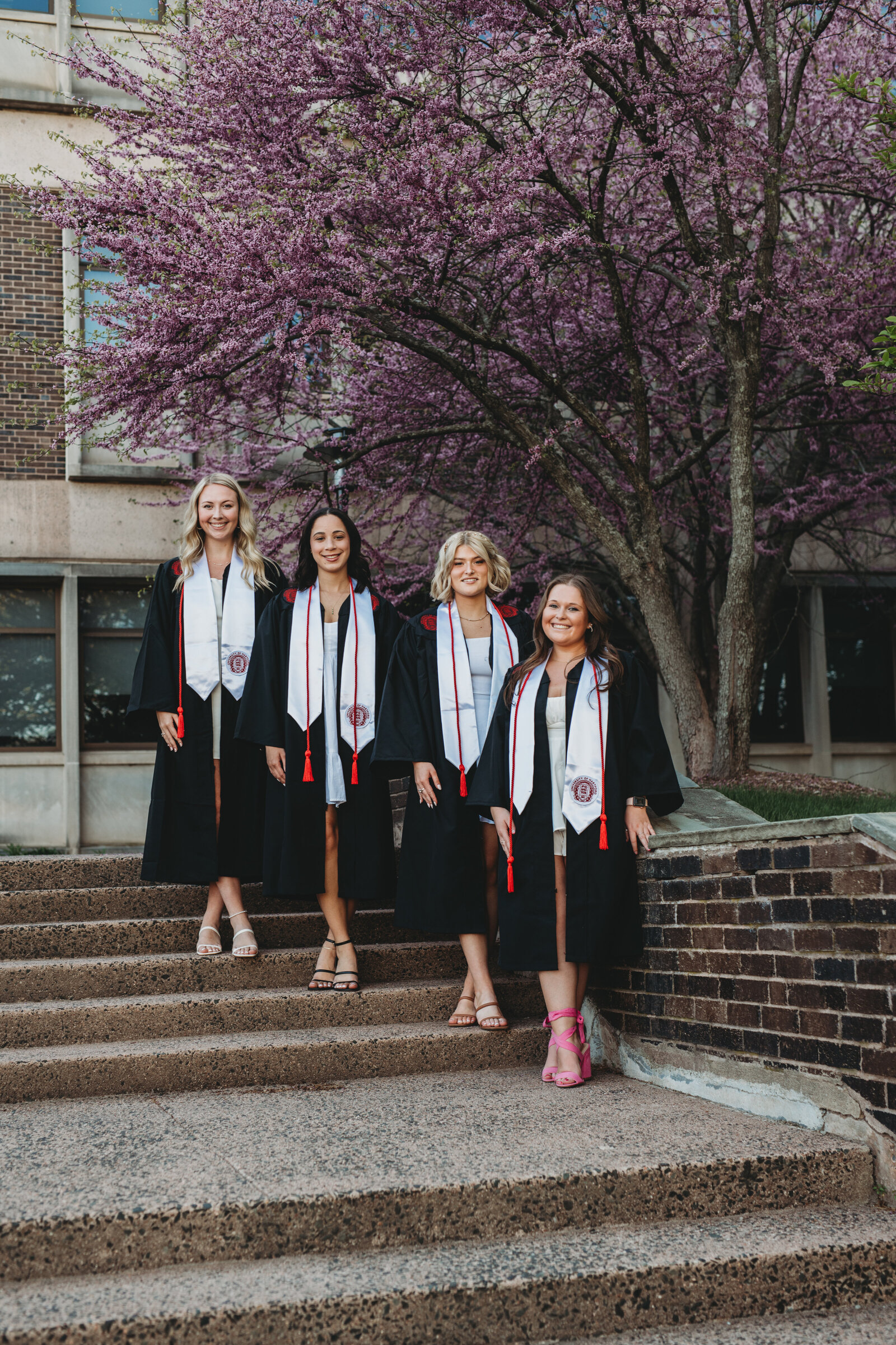 Four University of Hartford graduates in black caps and gowns pose on campus steps beneath a blooming cherry blossom tree, celebrating their achievement. Captured during a Connecticut college graduation photography session by Nicole Mele Photography, highlighting friendship and success.