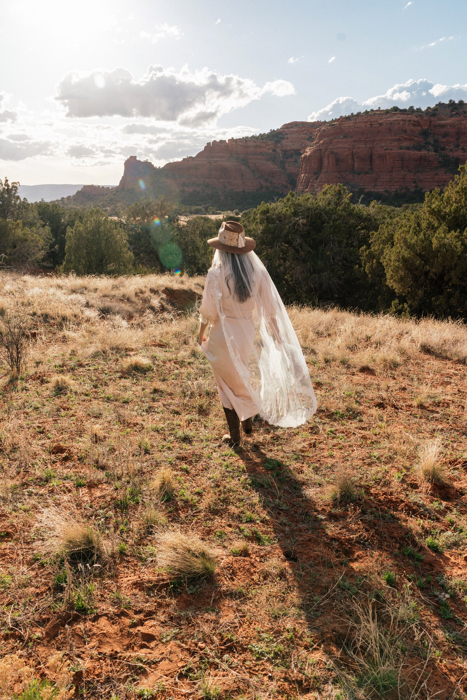 Bride in a vintage western dress and lace veil walking across the red rock terrain at Bear Mountain in Sedona, Arizona, with dramatic light and desert cliffs in the background.