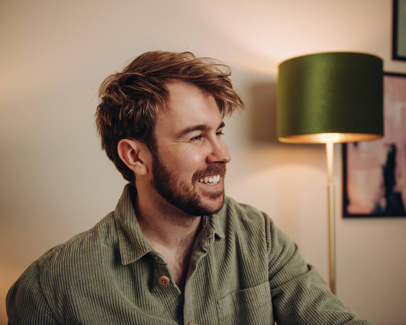 Tom Keenan smiling indoors, seated beside a green lamp.