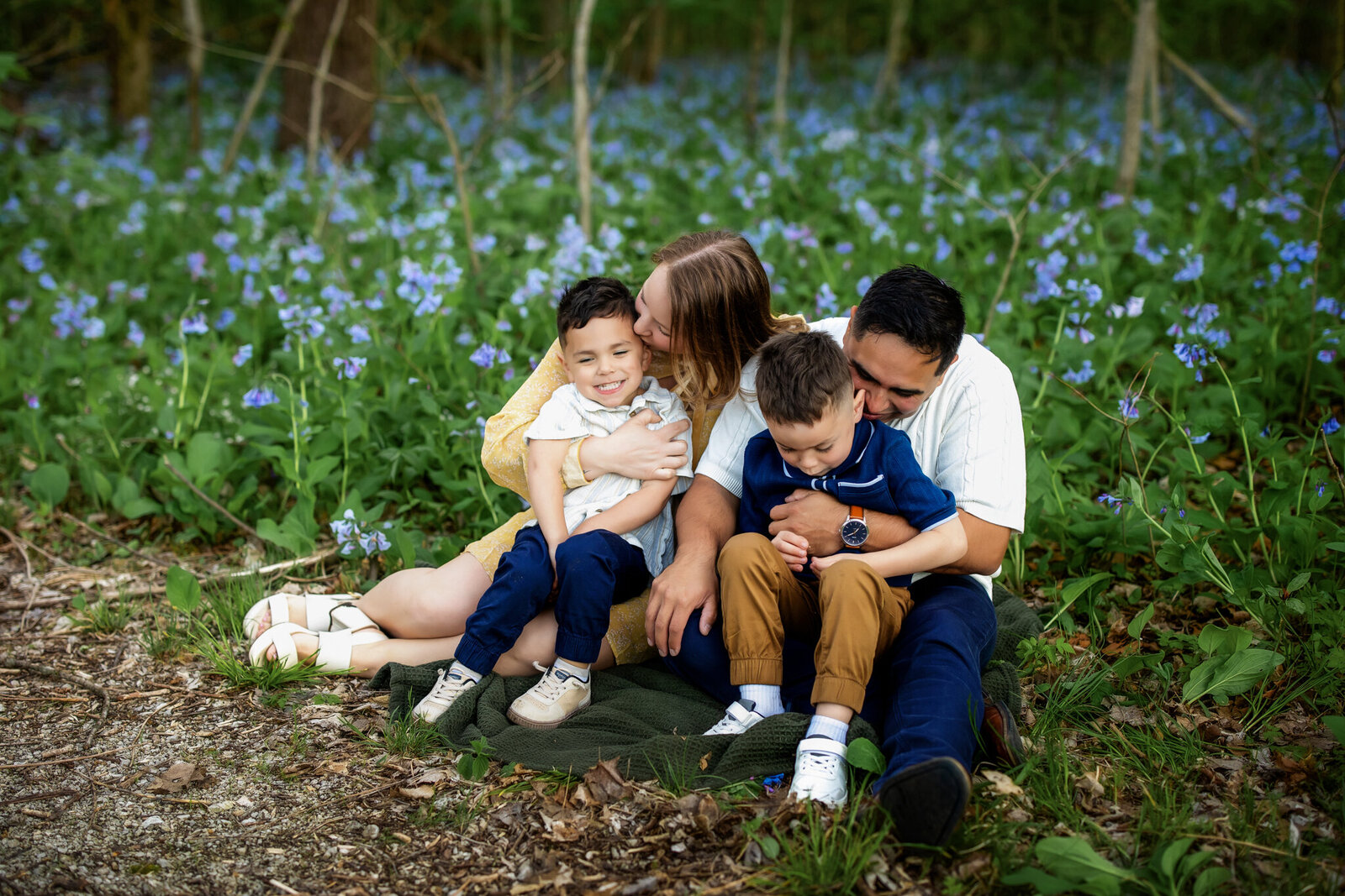 A Bloomington family photographer captures a family of four on a blanket in a forest clearing with blue wildflowers, the parents lovingly embracing their two young boys as they smile and laugh—outdoor portraits full of joy.