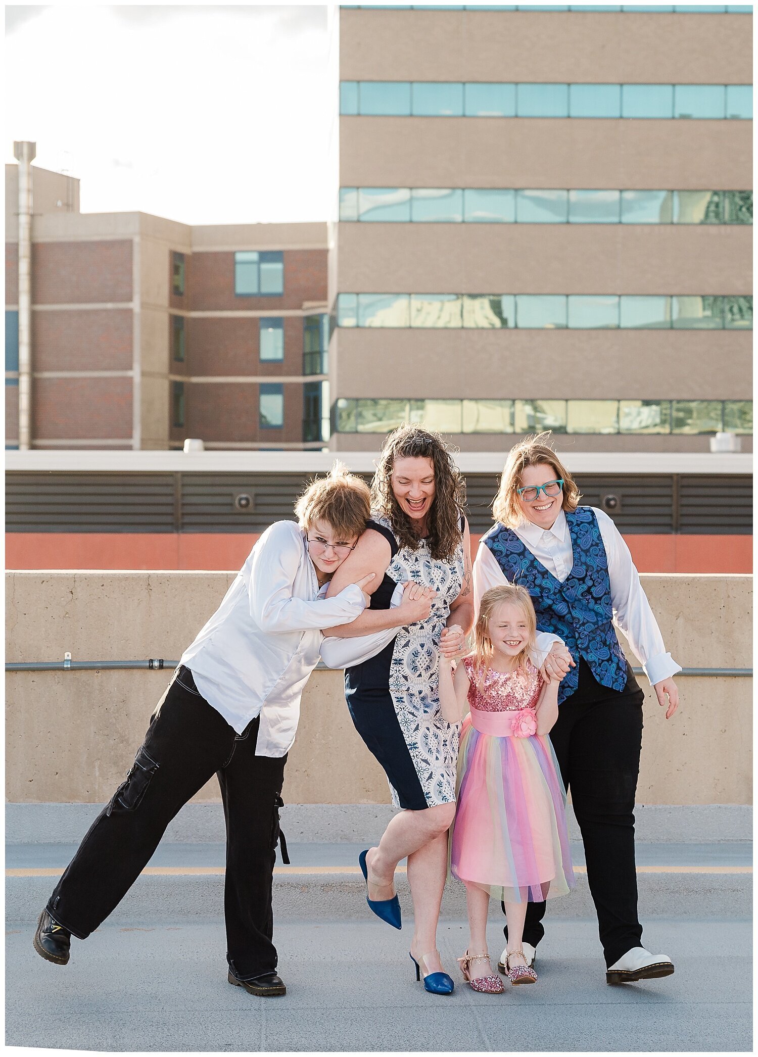 Two moms and 2 kids crash into each other in a big group hug on top of a rooftop in Denver with views of tall buildings behind them