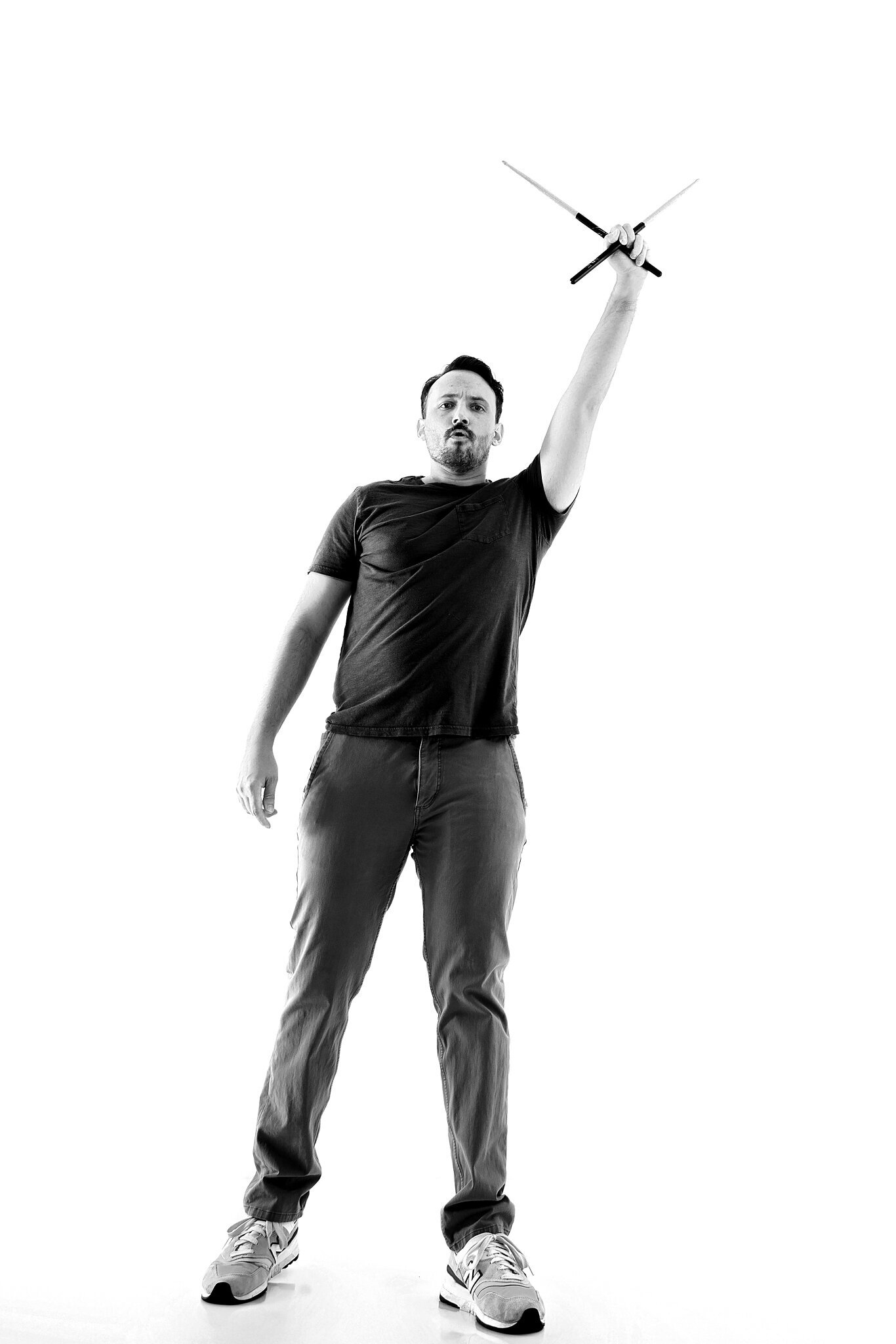 A man stands confidently, holding drumsticks high above his head with one hand, looking forward. Captured by a Jacksonville band photographer, the black and white image features a plain white background.