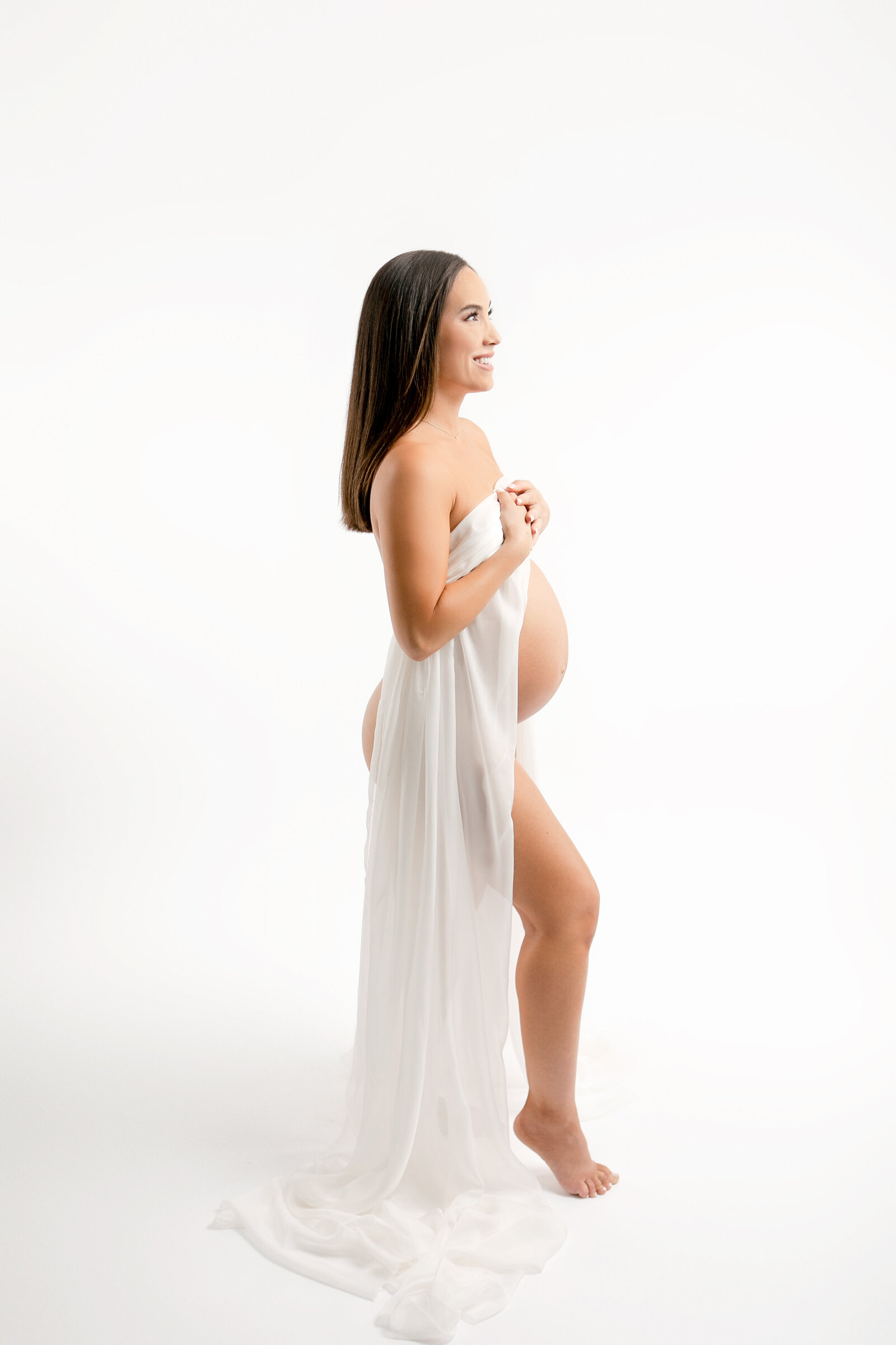 pregnant woman draped in a sheer white silk. all white backdrop, high key lighting, artsy, timeless, classic maternity photos in fresno, clovis, california