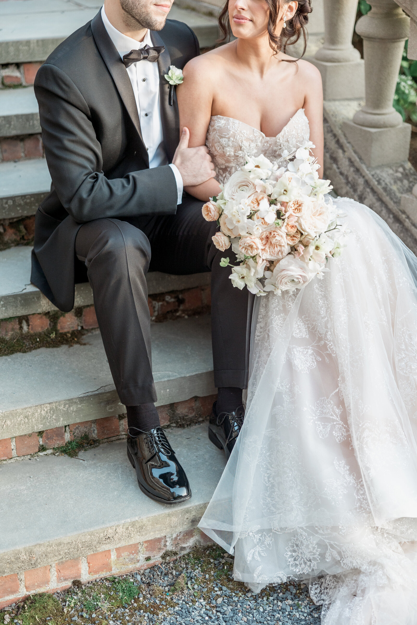 A couple cuddle on the steps of Blithewold Mansion. the bride has a blush and peach bouquet from Stoneblossom Florals in her hand.