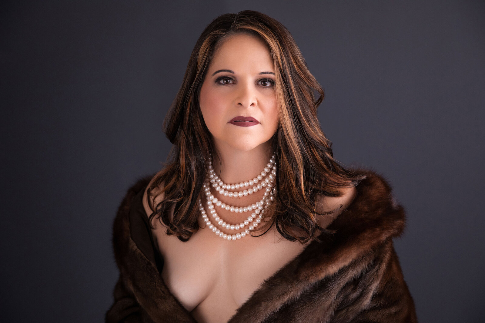 Woman posing in fur coat and pearls against simple gray background in Baton Rouge boudoir studio.