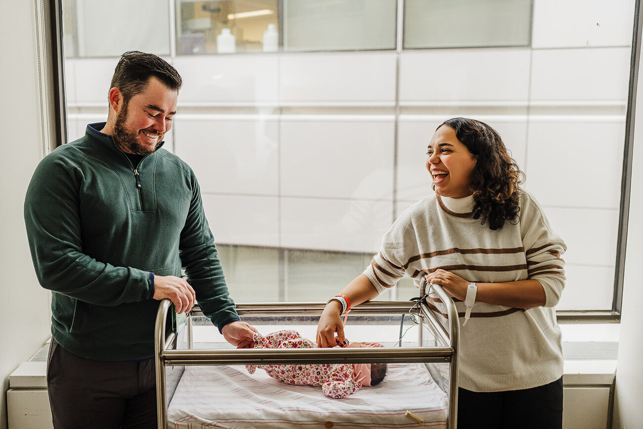 mom and dad laugh together standing over hospital bassinet during fresh 48 newborn photos