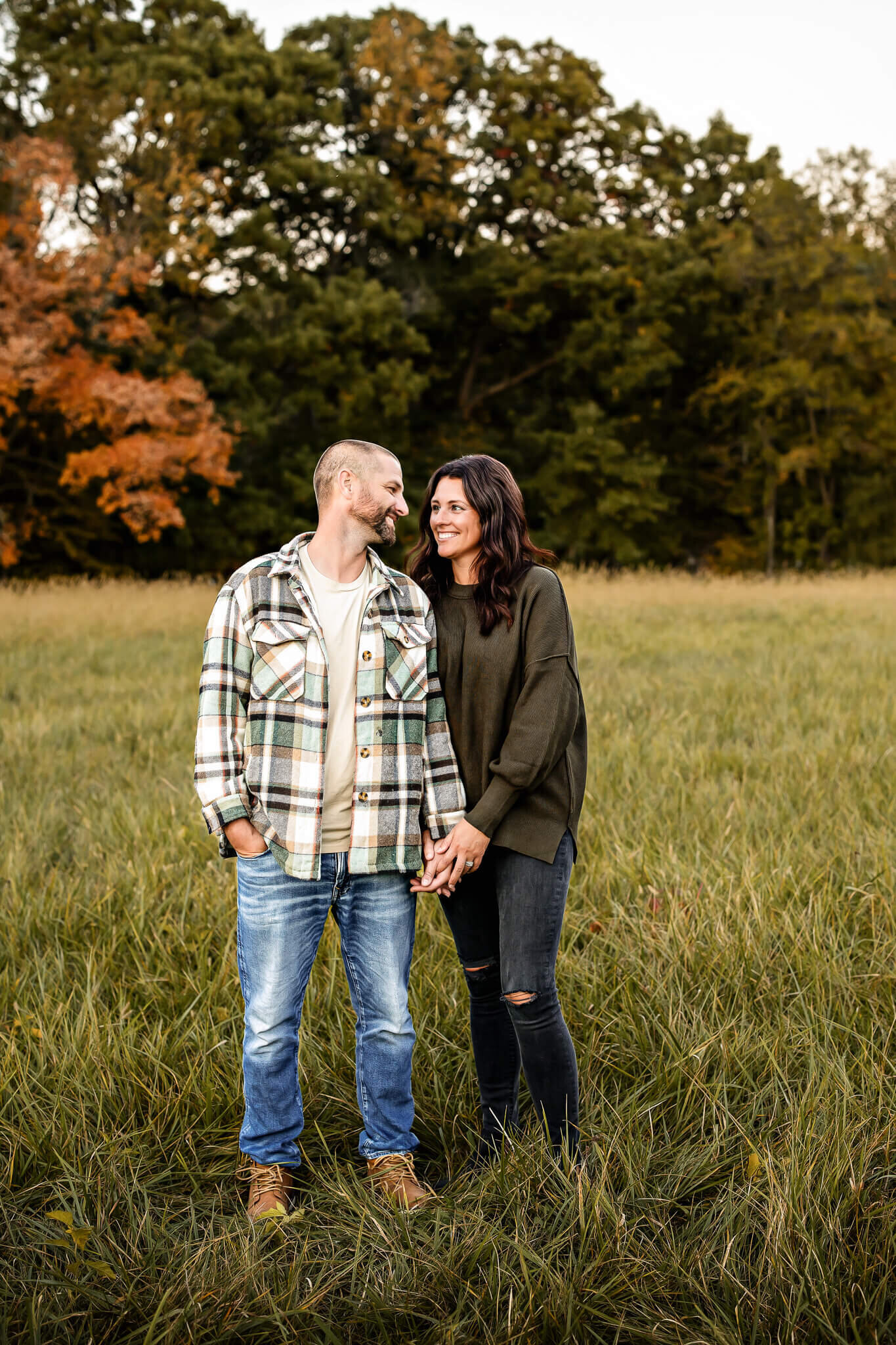 In a grassy field, a couple shares an affectionate glance while holding hands. The man dons a plaid jacket and jeans, the woman a dark sweater. Behind them, trees display vibrant green and autumnal hues, capturing the essence of outdoor portraits by a Bloomington family photographer.