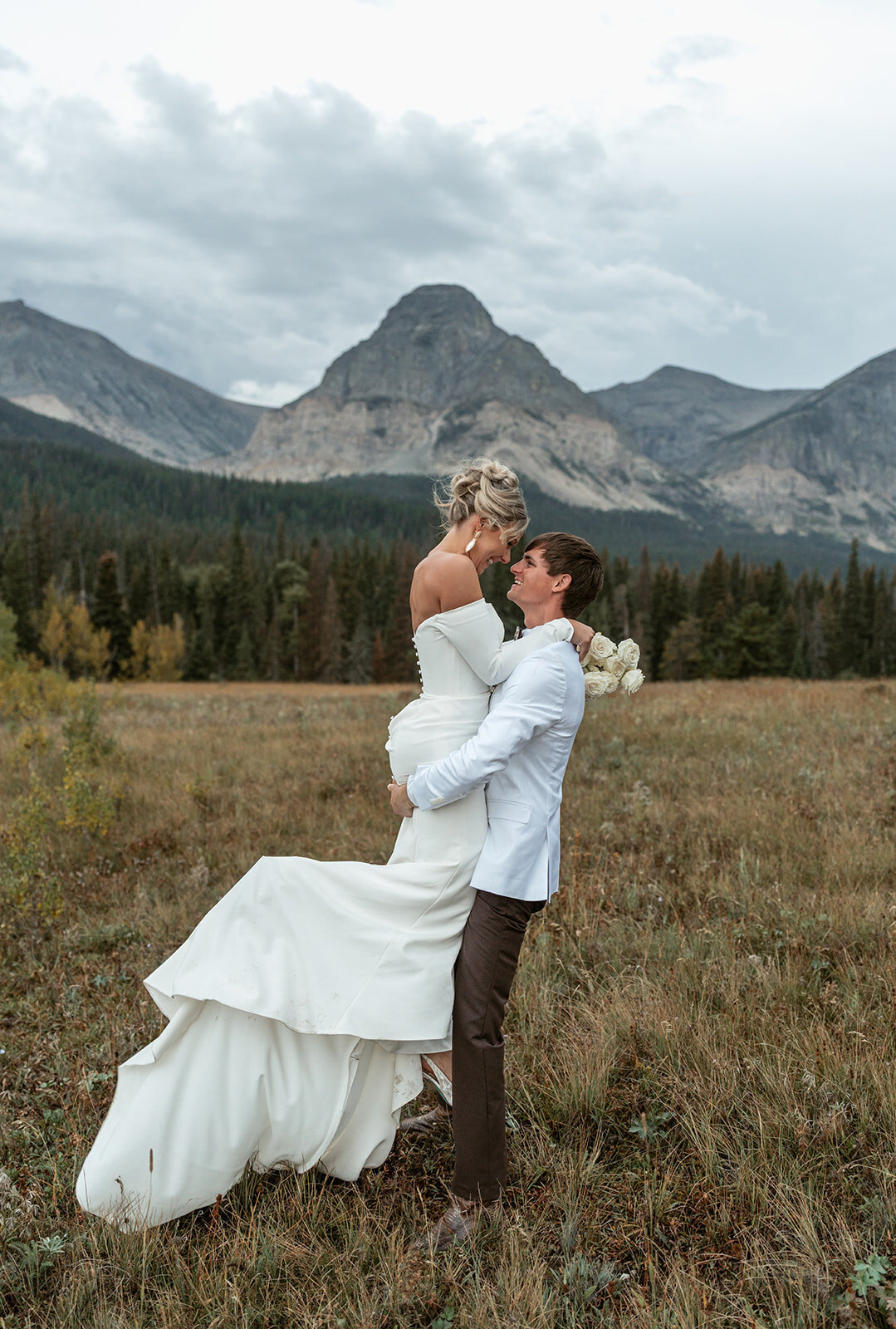 A groom lifts his bride in a grassy Montana meadow surrounded by rugged mountain peaks and pine forests during their Glacier National Park elopement, captured by Sydney Breann Photography.