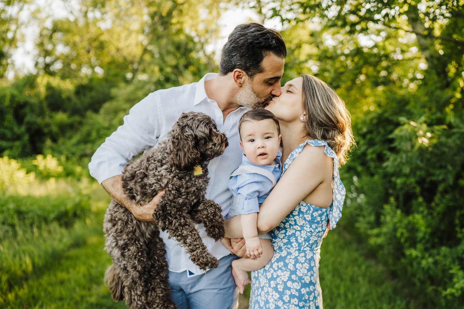 family kisses while holding baby boy and mini doodle
