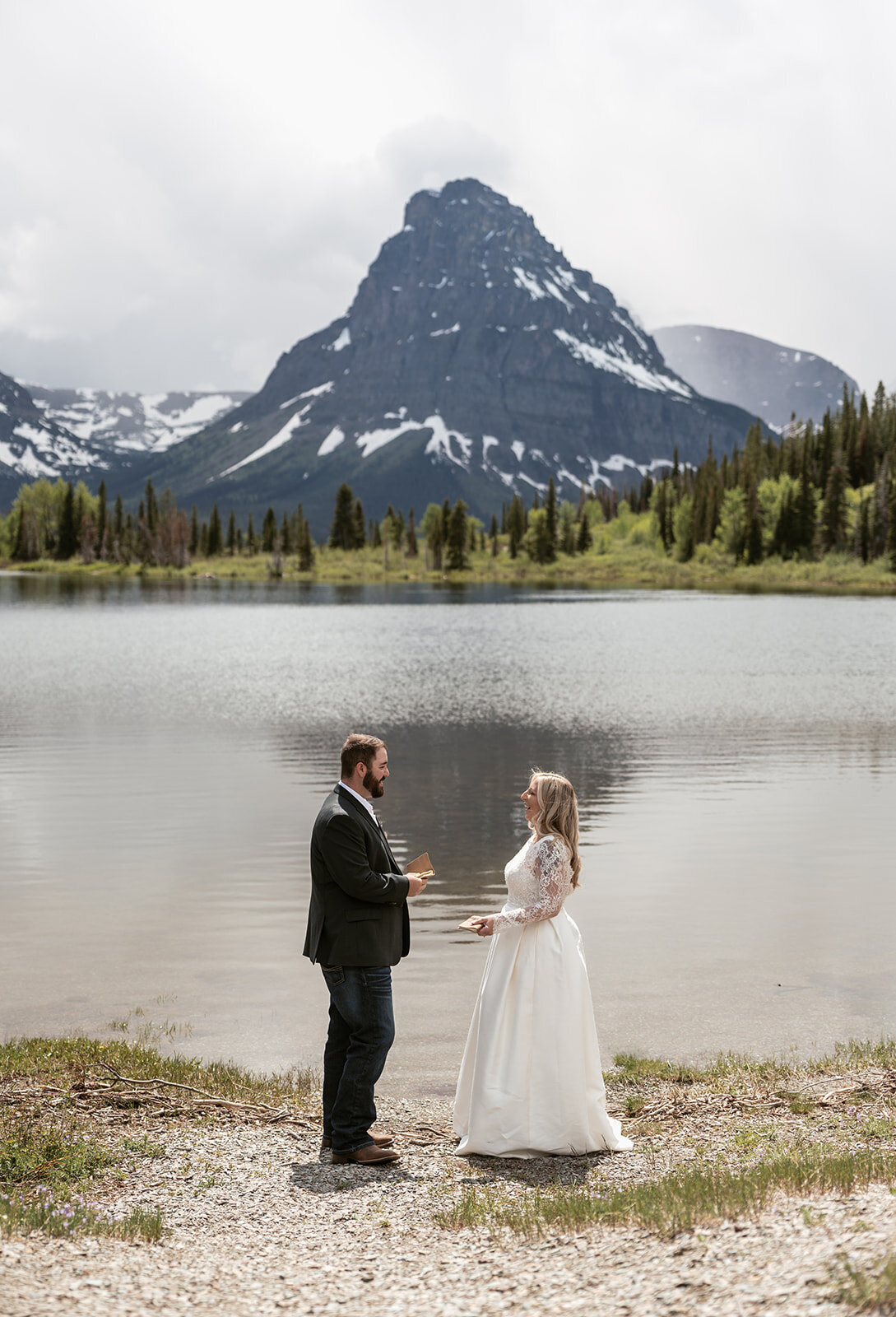 A bride and groom exchange vows beside a calm alpine lake surrounded by pine trees and snow-capped peaks in Glacier National Park, captured by Sydney Breann Photography during their intimate Montana elopement.