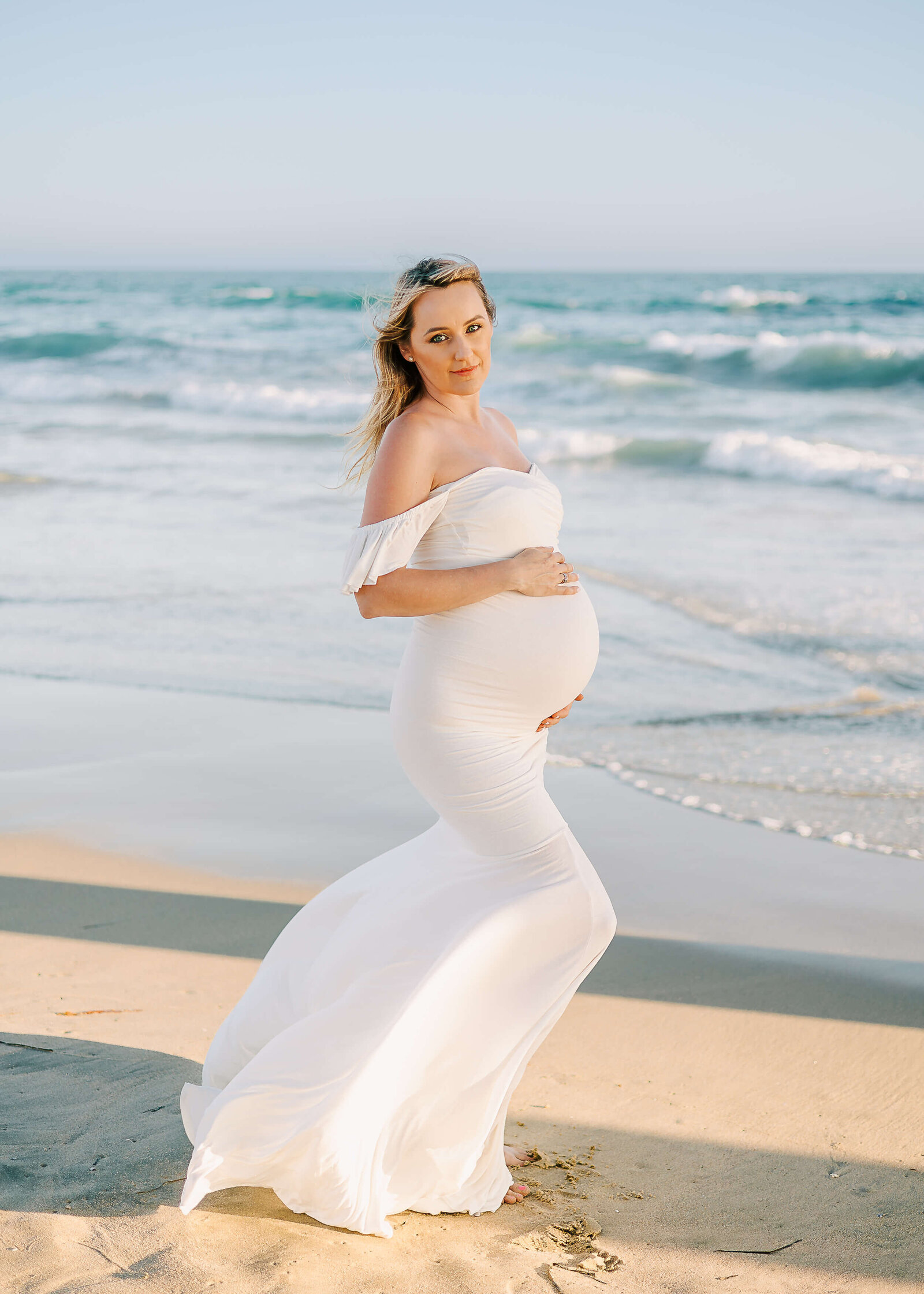 Gorgeous expectant mom standing holding baby bump on beach in Orange County, CA by Ashley Nicole.