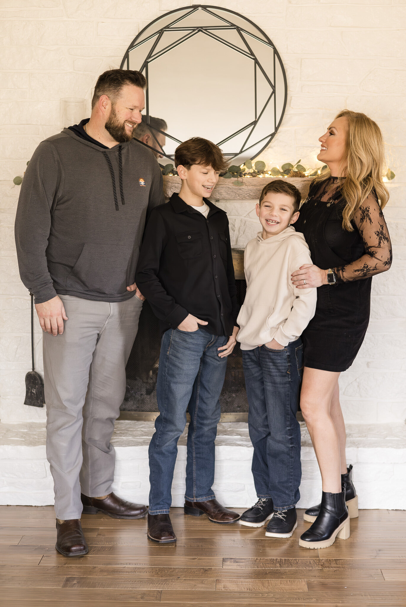 A husband, wife, and two sons standing in front of a fireplace, all looking at each other laughing.  Dad has dark hair and beard, wearing a dark gray hoodie, light gray pants, and black shoes.  Older son has dark hair, wearing a black shirt, blue jeans, and wearing dark boots.  Younger son has a light tan long sleeve shirt, dark hair, and blue jeans.  Mom is wearing a black dress with lace sleeves, black boots, and has long blonde hair.