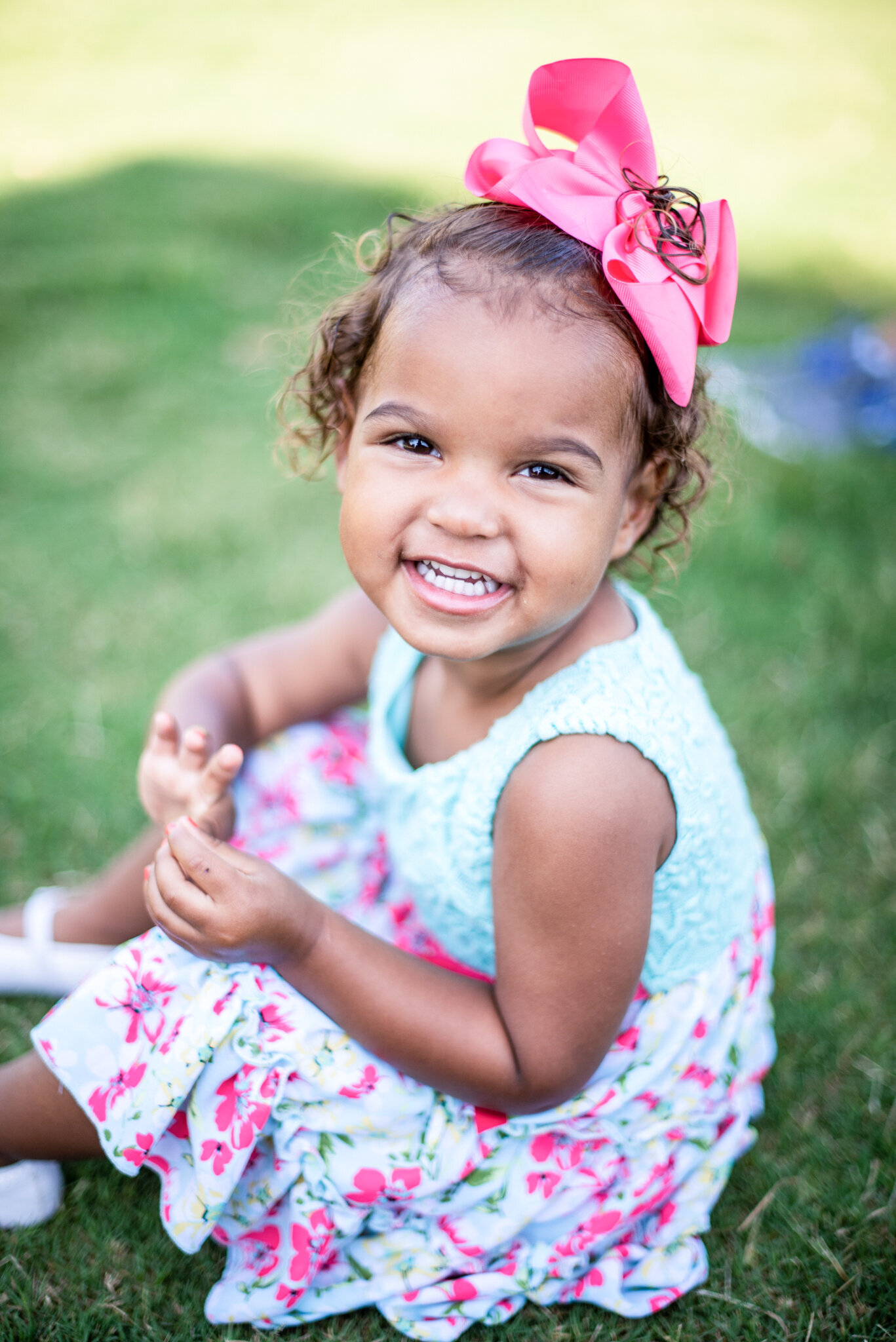 young girl in a white and pink floral dress with a large pink bow on her head sitting in the grass smiling at the camera photographed by Millz Photography in Greenville, SC